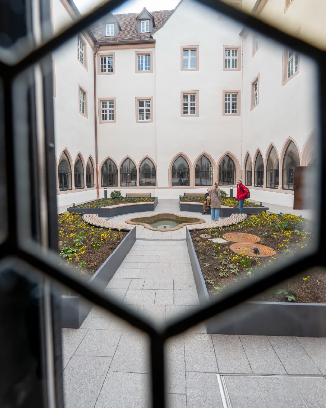 Dieser Alternativtext wurde KI-generiert: Ein Innenhof mit einem Garten ist durch einen ungewöhnlich geformten Rahmen zu sehen. Mehrere Personen stehen im Hof und betrachten die Bepflanzung.