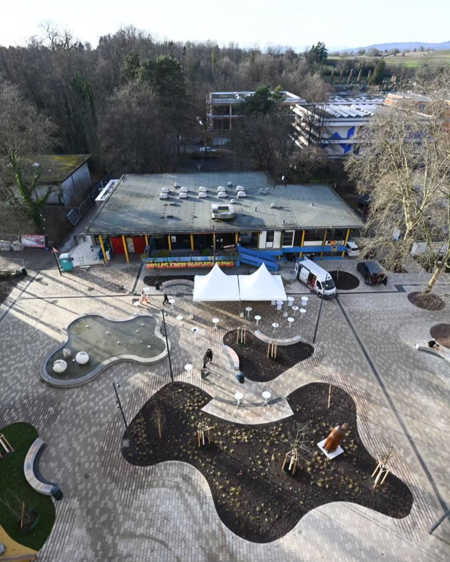 Dieser Alternativtext wurde KI-generiert: Eine Vogelperspektive zeigt Gebäude mit einem farbenfrohen äußeren Erscheinungsbild und einem umgebenden gepflasterten Bereich mit Bäumen und mehreren weißen Zelten. Das Bild ist farbig und zeigt eine Außenansicht.