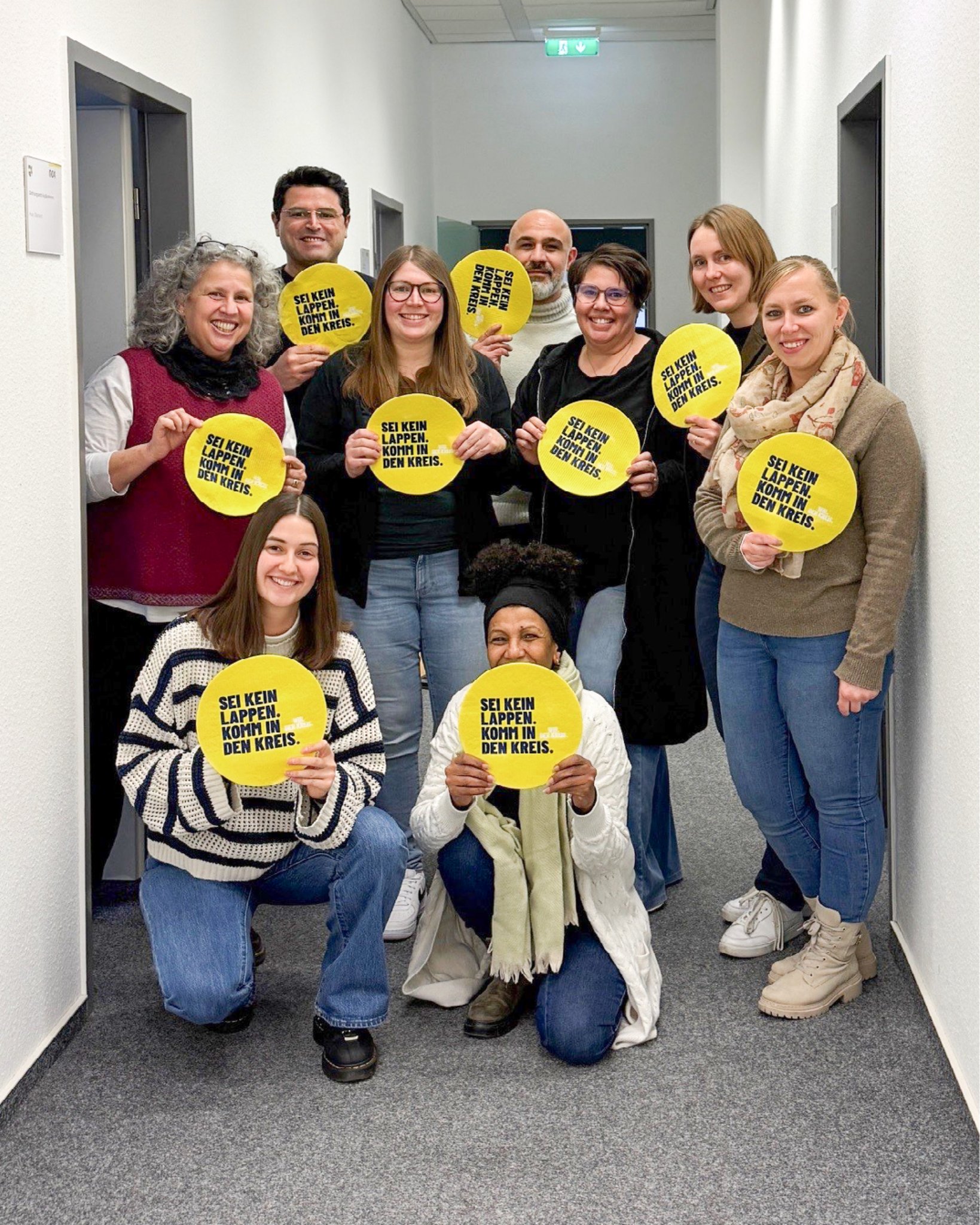 Dieser Alternativtext wurde KI-generiert: Eine Gruppe von Menschen hält Schilder mit Text in den Händen. Die Aufnahme ist farbig und zeigt eine helle Innenraumgestaltung.