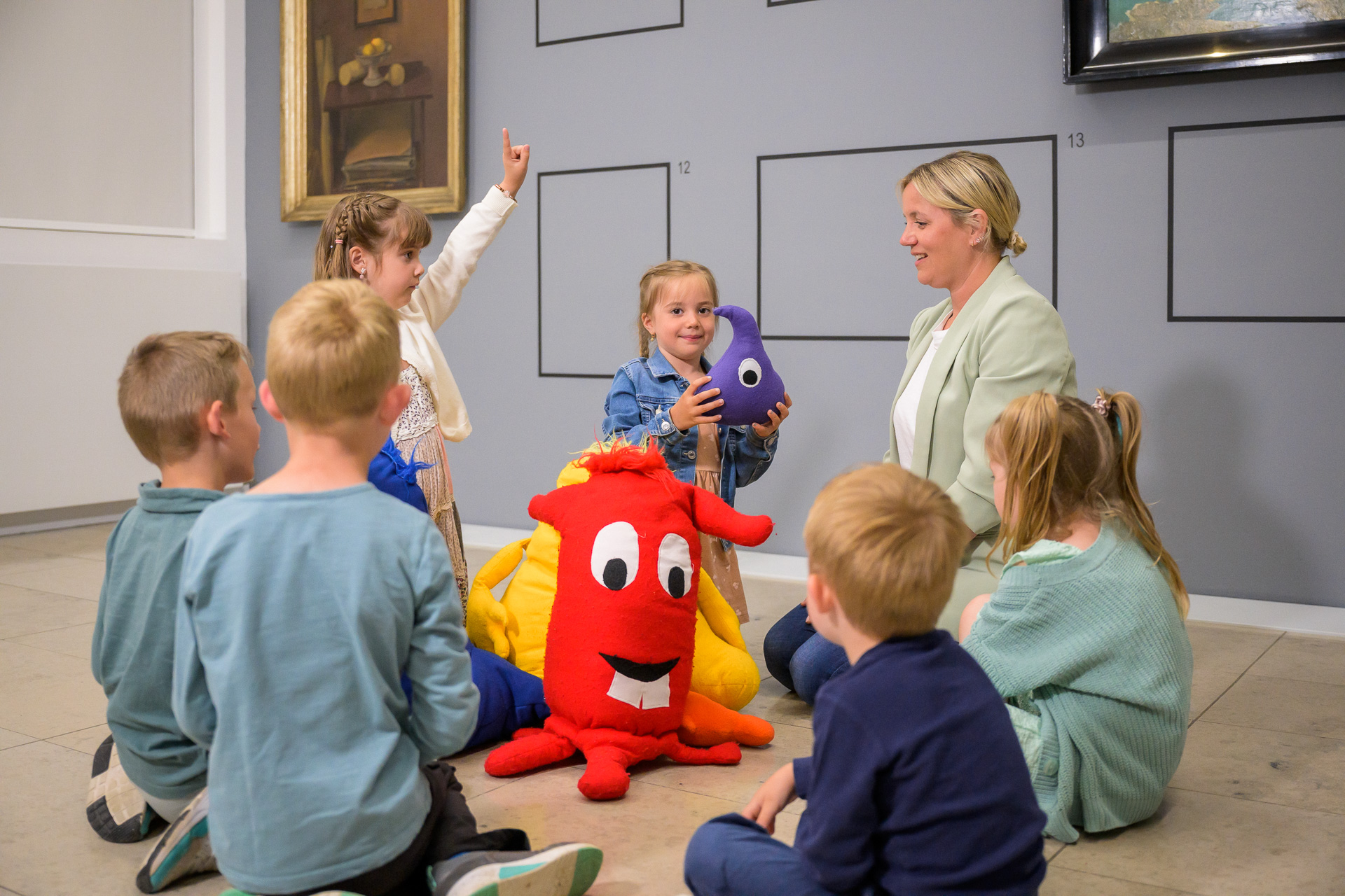 Mehrere Kinder sitzen im Kreis auf dem Boden eines Raums mit Bildern an der Wand. Ein stehendes Kind zeigt nach oben, ein anderes hält ein lila Plüschtier. In der Mitte sitzt ein großer roter Plüschmonster, daneben eine Erwachsene, die die Gruppe ansieht. (KI-Alternativtext)