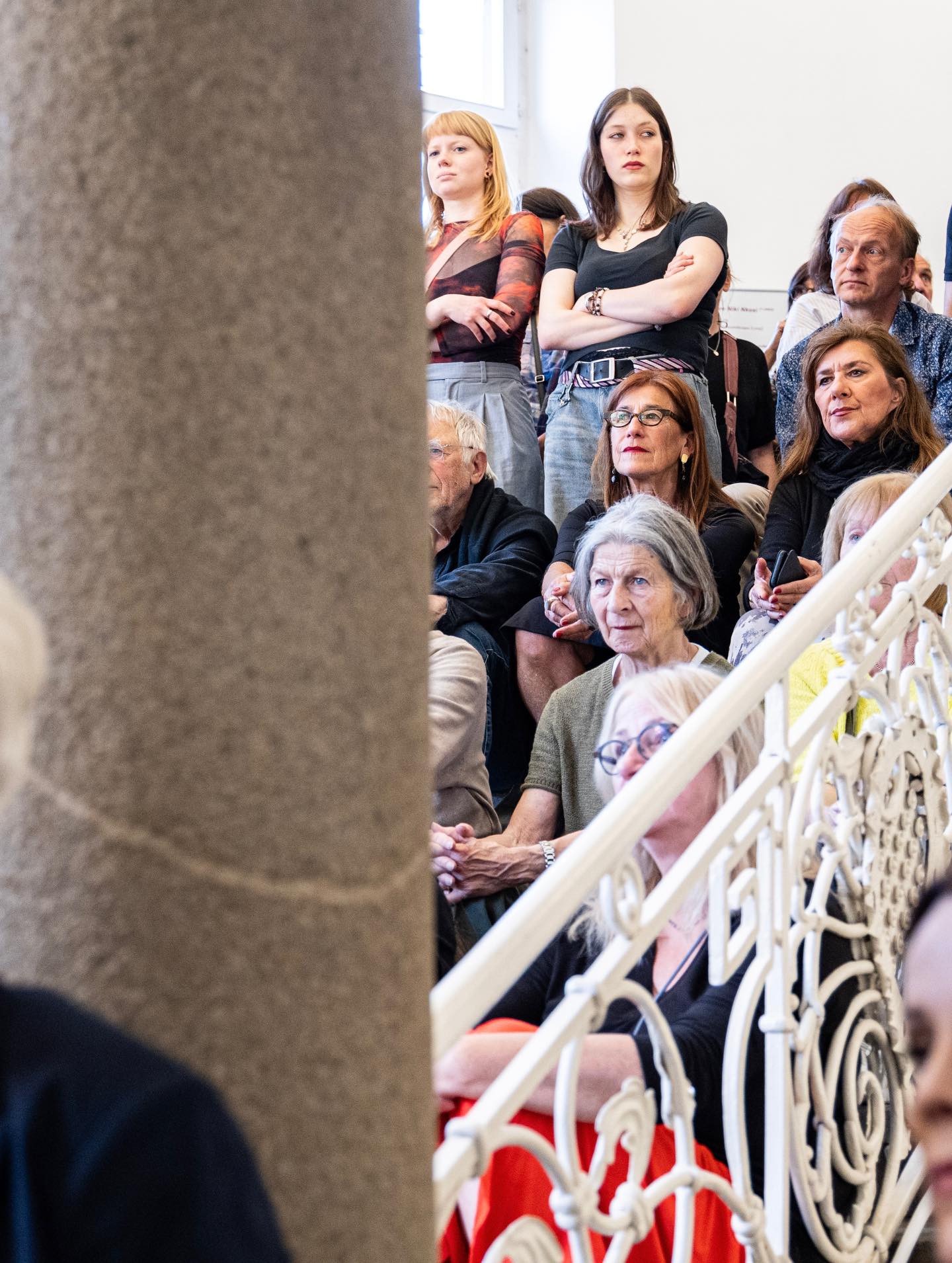 Dieser Alternativtext wurde KI-generiert: Eine Gruppe von Menschen sitzt und steht auf einer Empore oder Treppe. Der Blick ist auf die Personen gerichtet, die sich inmitten einer verzierenden Metallstruktur befinden.