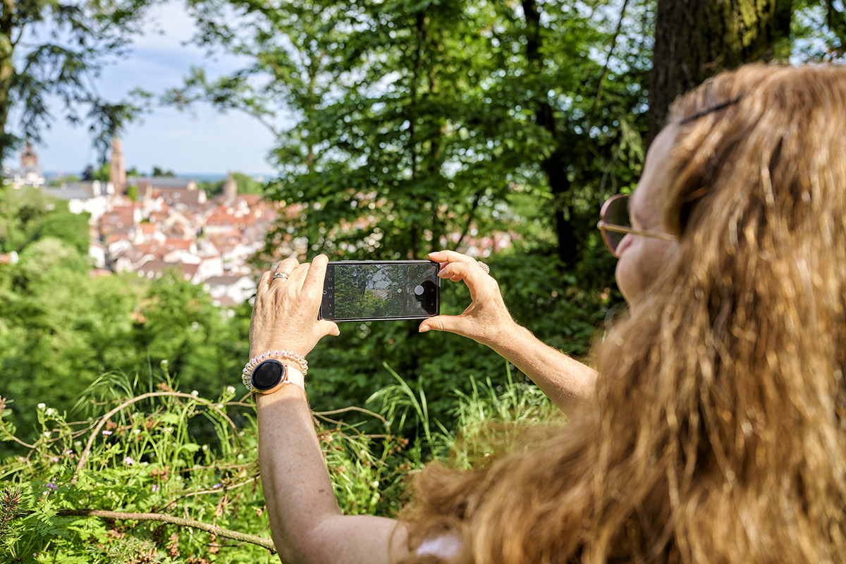 Eine Person mit langen, lockigen Haaren und Sonnenbrille macht mit einem Smartphone ein Foto von einer Stadtansicht mit roten Dächern und Kirchtürmen, umgeben von grünen Bäumen. (KI-Alternativtext)