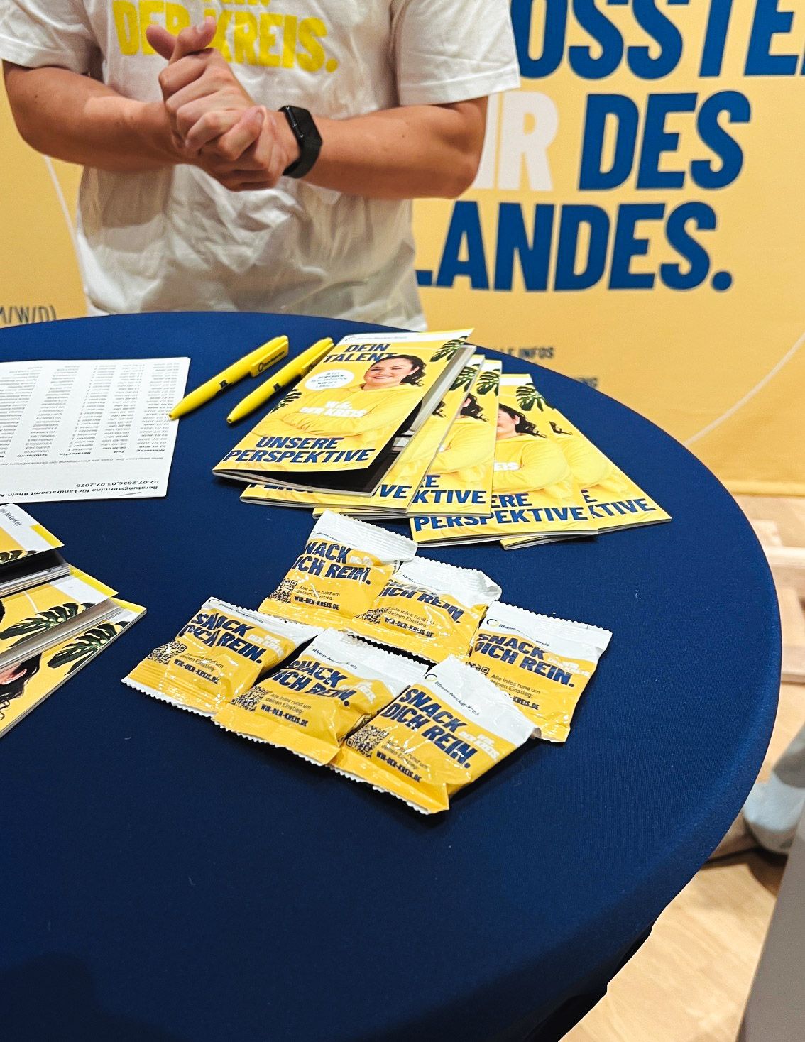 Auf einem blauen Tisch liegen gelbe Broschüren mit den Aufschriften „Dein Talent“ und „Unsere Perspektive“ sowie kleine Snack-Packungen mit dem Text „Snack dich rein“. Daneben liegen gelbe Stifte und ein Zettel. Im Hintergrund steht eine Person in einem weißen T-Shirt vor einem gelben Banner mit dem Text „des Landes“ und reibt die Hände. (KI-Alternativtext)