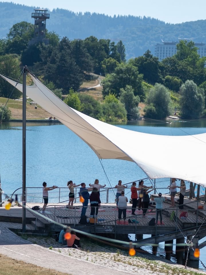 Mehrere Personen stehen auf einer Holzplattform am Ufer eines Sees unter einem großen weißen Sonnensegel und führen synchron eine Übung mit ausgestreckten Armen aus. Im Hintergrund sind bewaldete Hügel, ein Aussichtsturm und ein Gebäude zu sehen. (KI-Alternativtext)