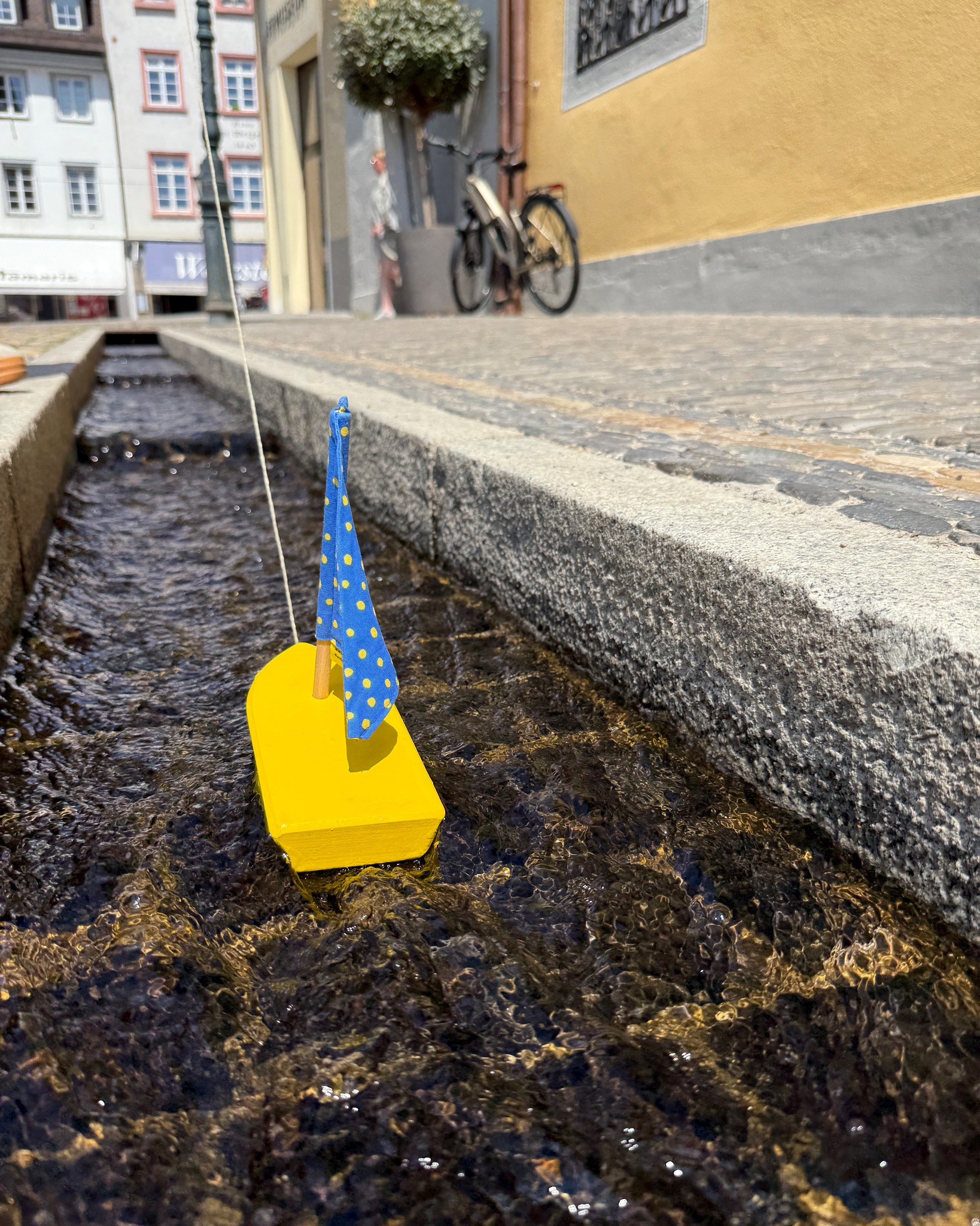 Ein gelbes Spielzeugboot mit blauem Segel und gelben Punkten treibt in einem schmalen, steinernen Wasserlauf. Im unscharfen Hintergrund sind städtische Gebäude, ein geparktes Fahrrad und eine Person zu erkennen. (KI-Alternativtext)