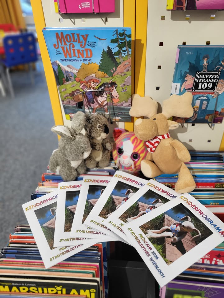 Dieser Alternativtext wurde KI-generiert: Eine Ansammlung von Büchern und Spielzeug steht auf einem Regal. Darunter befinden sich auch einige Flyer und ein Teddybär.