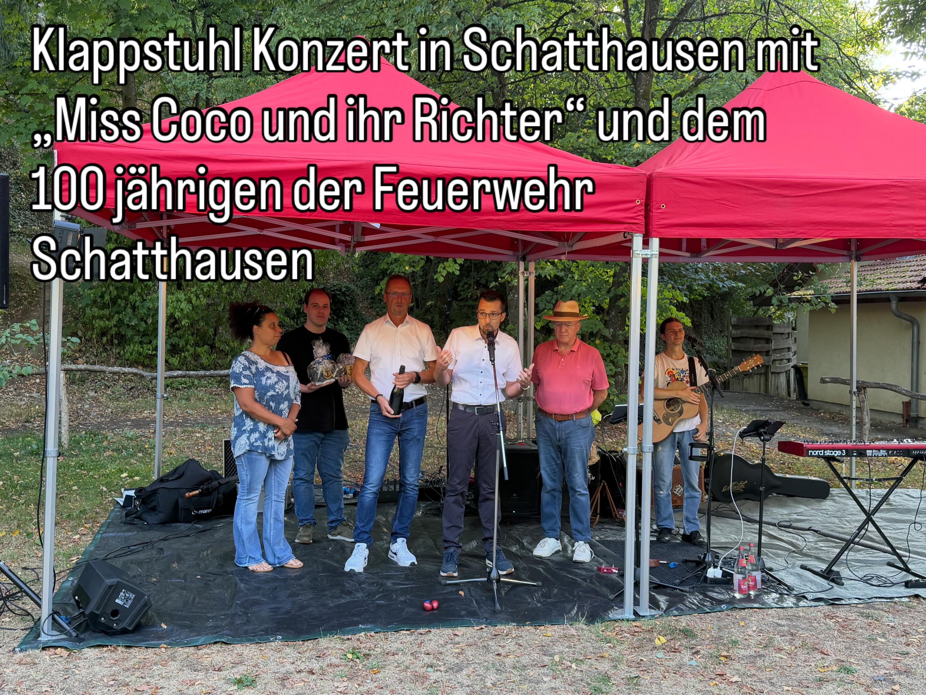 Über dem Bild steht der Text: „Klappstuhl Konzert in Schatthausen mit ‚Miss Coco und ihr Richter‘ und dem 100 jährigen der Feuerwehr Schatthausen“. Auf einer Bühne unter roten Zelten stehen sechs Personen. Eine Person spricht in ein Mikrofon, eine weitere spielt Gitarre, während andere Personen Geschenke oder eine Flasche halten. (KI-Alternativtext)