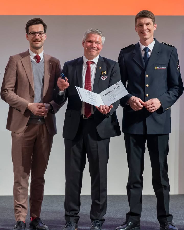 Dieser Alternativtext wurde KI-generiert: Drei Personen in formeller Kleidung stehen nebeneinander und betrachten ein Dokument. Im Hintergrund ist ein orangefarbener Farbverlauf zu sehen.