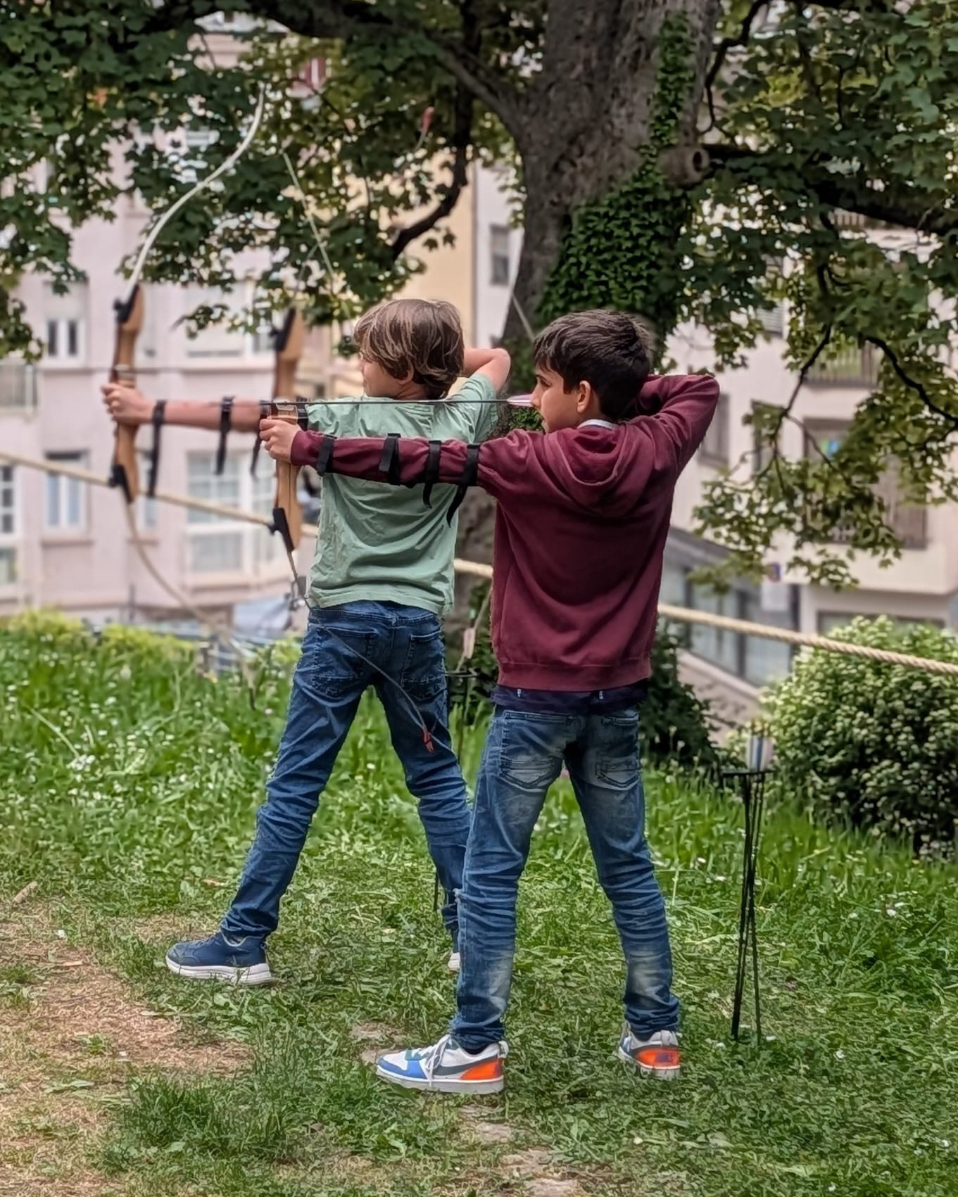Zwei Kinder stehen im Freien auf einer Wiese und zielen mit Bögen auf ein Ziel. Im Hintergrund sind Bäume und ein Gebäude zu sehen. (KI-Alternativtext)