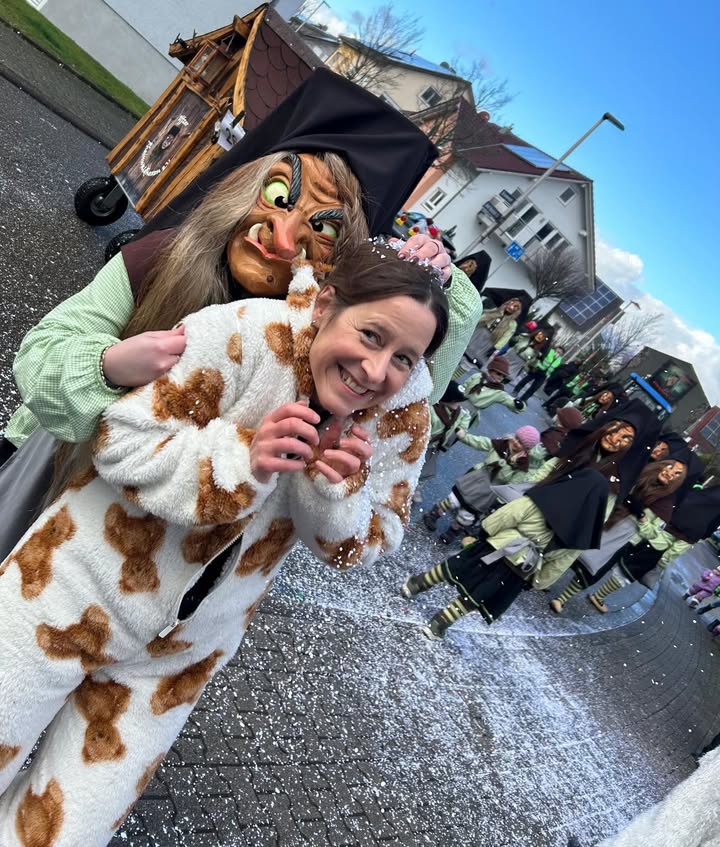 Dieser Alternativtext wurde KI-generiert: Mehrere Personen in Kostümen stehen auf einer Straße und lächeln in die Kamera. Im Hintergrund sind weitere verkleidete Gestalten und Häuser zu sehen.