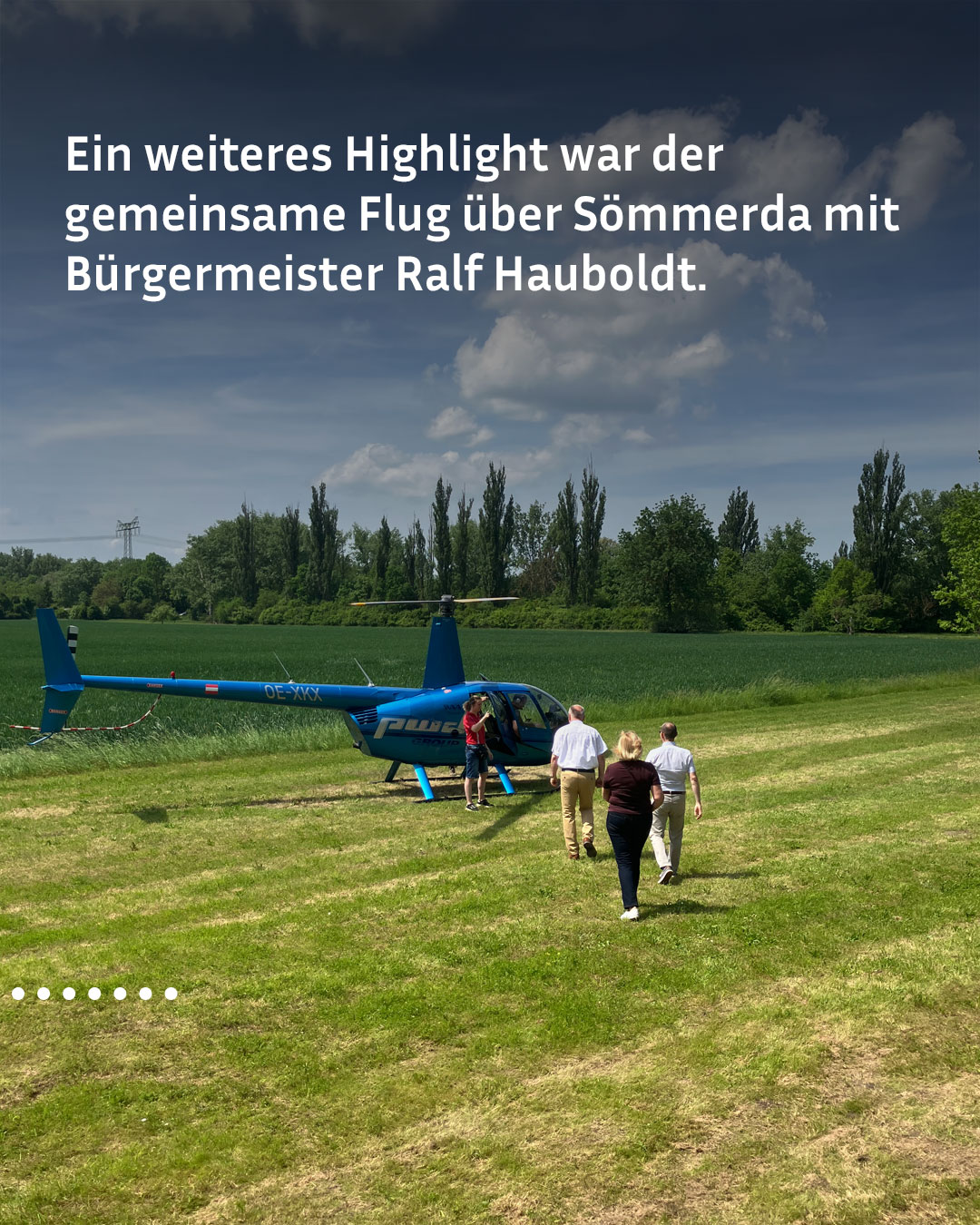 Ein blauer Hubschrauber steht auf einer Wiese, mehrere Personen gehen darauf zu. Im Hintergrund sind Bäume und ein bewölkter Himmel zu sehen. Der Text im Bild erwähnt einen gemeinsamen Flug über Sömmerda mit Bürgermeister Ralf Hauboldt. (KI-Alternativtext)
