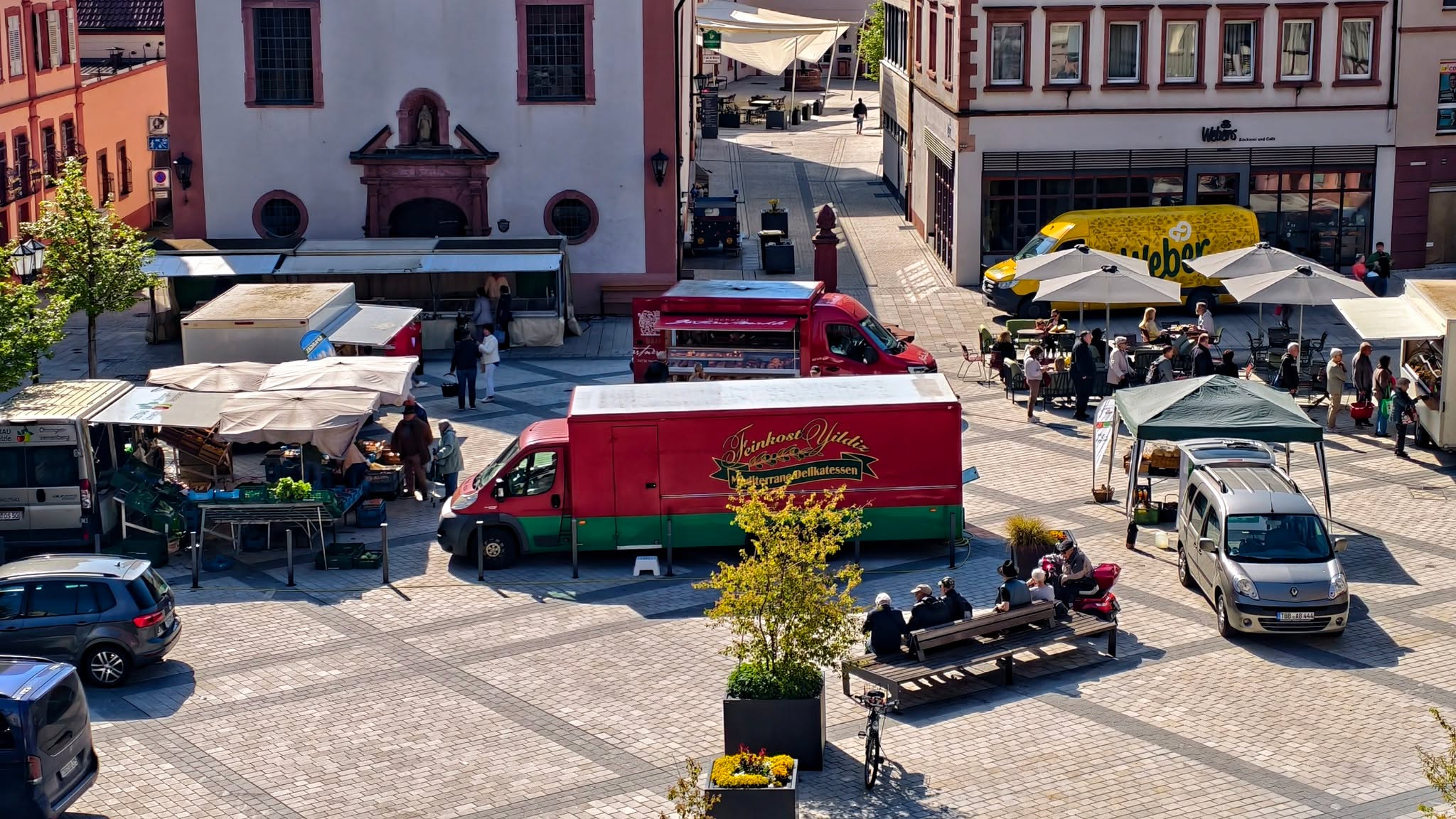 Dieser Alternativtext wurde KI-generiert: Ein belebter Platz mit Marktständen und Sitzgelegenheiten. Mehrere Menschen sind zu sehen, die sich dort aufhalten oder einkaufen.