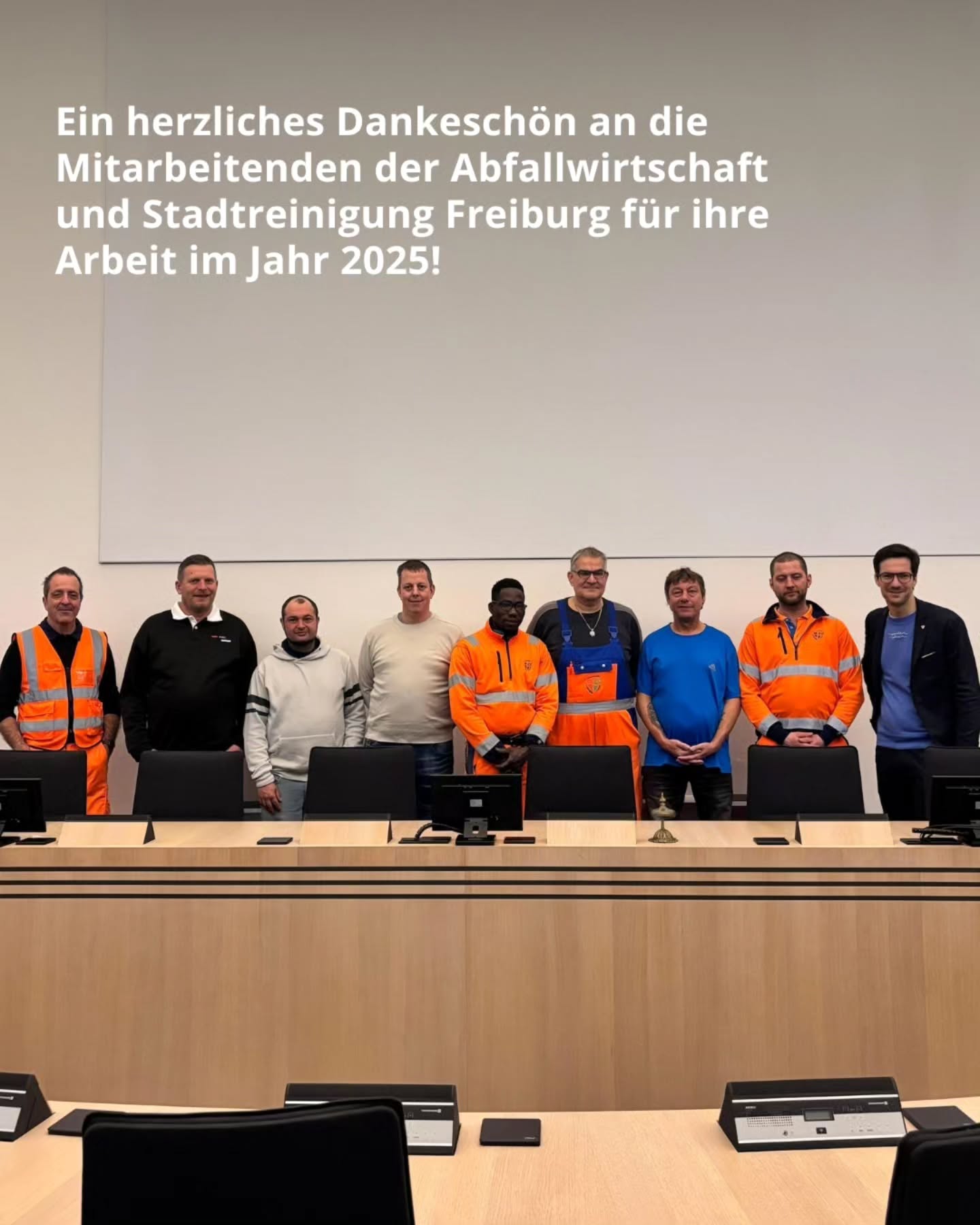 Dieser Alternativtext wurde KI-generiert: Ein Gruppenfoto von Personen in Arbeitskleidung vor einer Reihe von leeren Stühlen und einem Pult. Der Hintergrund besteht aus Holzpaneelen und einer Glasscheibe.