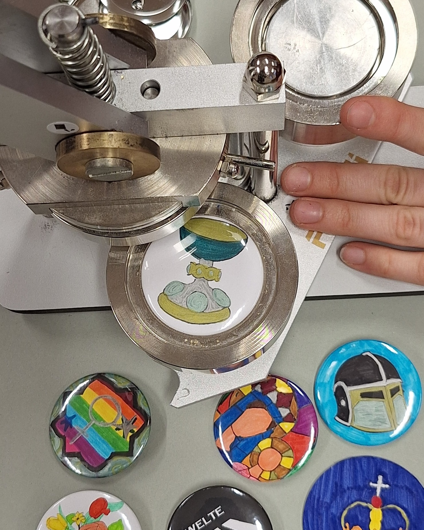 Dieser Alternativtext wurde KI-generiert: Ein metallisches Gerät wird verwendet, um einen runden Aufkleber zu erstellen, der ein buntes Bild zeigt. Mehrere fertige, runde Aufkleber liegen daneben.