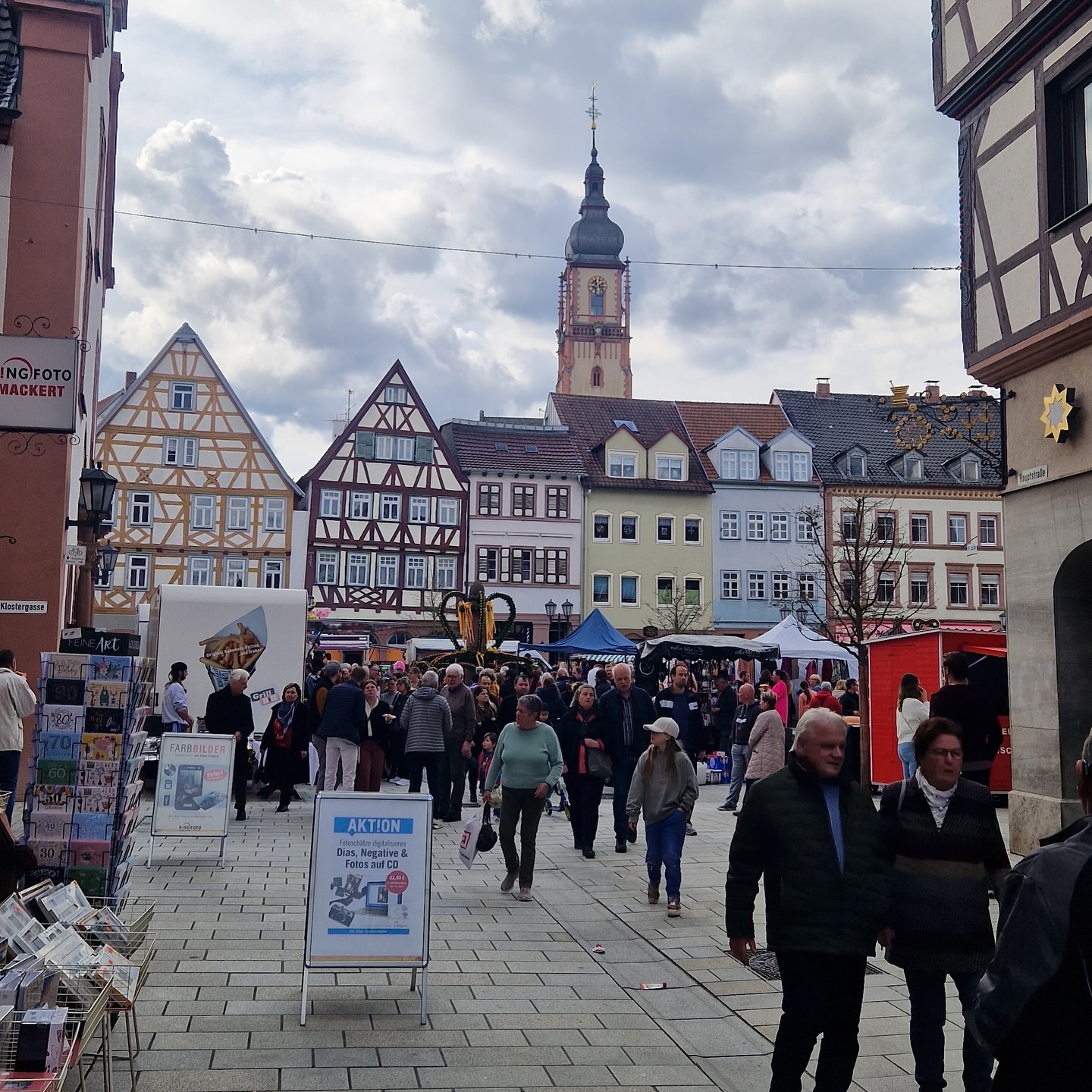 Menschen flanieren über einen Marktplatz mit Fachwerkhäusern und einem Kirchturm im Hintergrund. Verschiedene Marktstände sind aufgebaut.