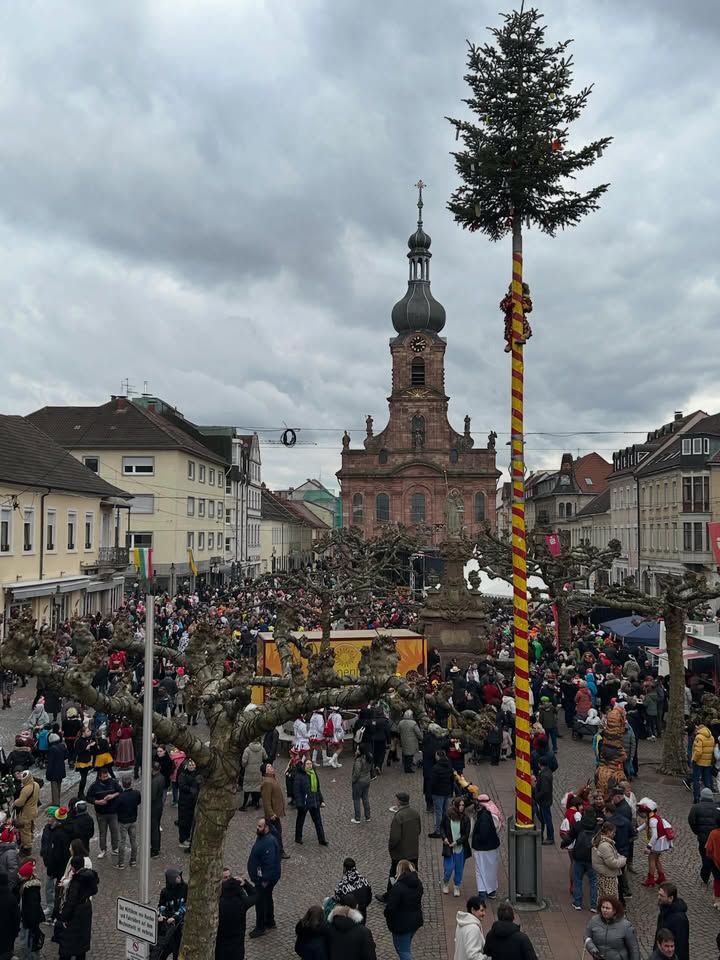 Dieser Alternativtext wurde KI-generiert: Ein hoher, schlanker Tannenbaum steht vor einem historischen Kirchturm und einer belebten Stadtansicht an einem bewölkten Tag. Im Vordergrund ist eine Menschenmenge auf einem Platz zu sehen.