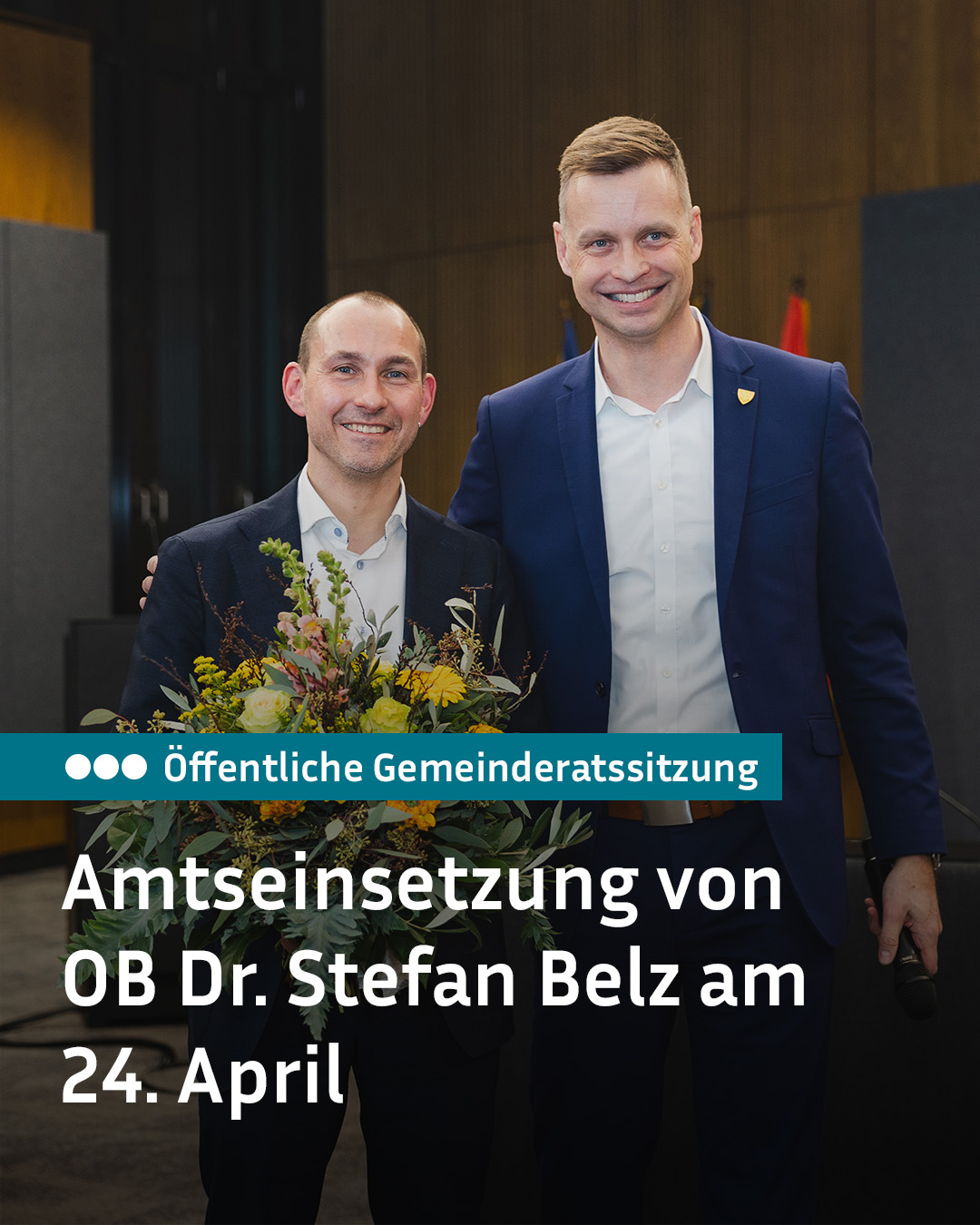 Dieser Alternativtext wurde KI-generiert: Zwei Personen mit Anzug stehen lächelnd nebeneinander und halten Blumen. Im Hintergrund ist ein Schild mit Text zu sehen.