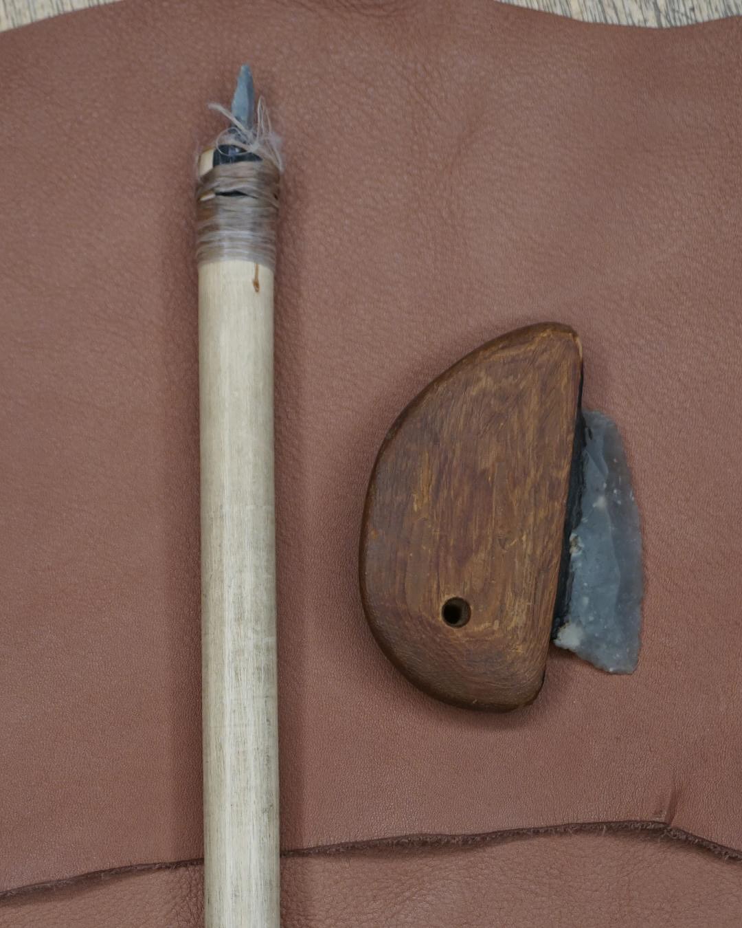 Ein hölzerner Speer mit steinerner Spitze, befestigt mit Schnur, liegt neben einem hölzernen Werkzeug mit eingesetztem Stein auf braunem Leder. (KI-Alternativtext)