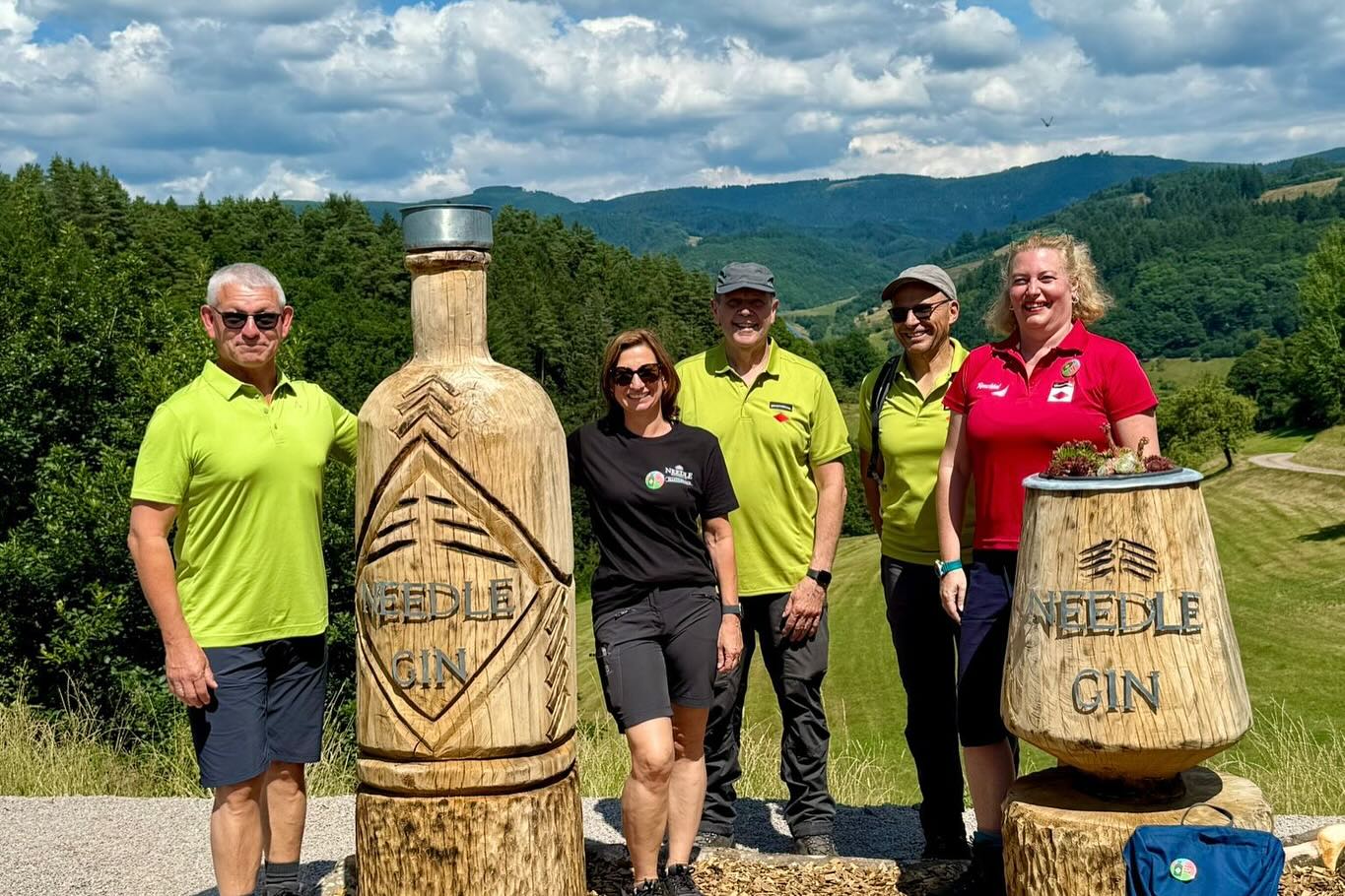 Im Freien posieren Personen neben einer Holzskulptur in Flaschenform und einer Fassskulptur mit dem Schriftzug "Needle Gin" vor einer hügeligen Landschaft.