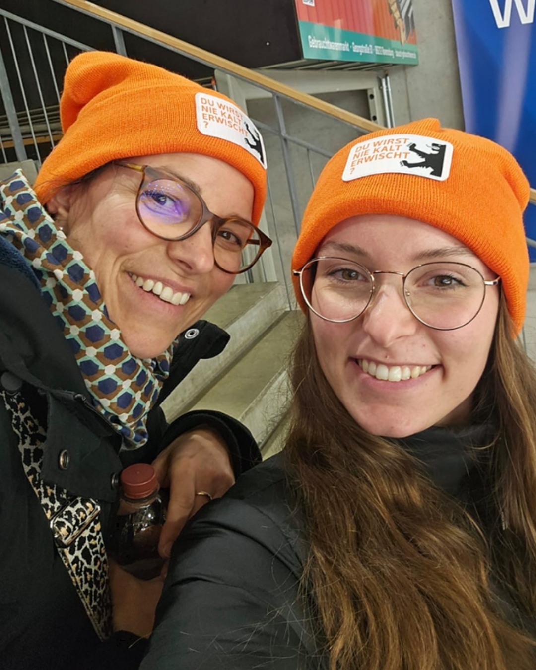 Dieser Alternativtext wurde KI-generiert: Zwei Personen mit orangefarbenen Strickmützen lächeln in die Kamera. Beide tragen Brillen und sind in Innenräumen zu sehen.