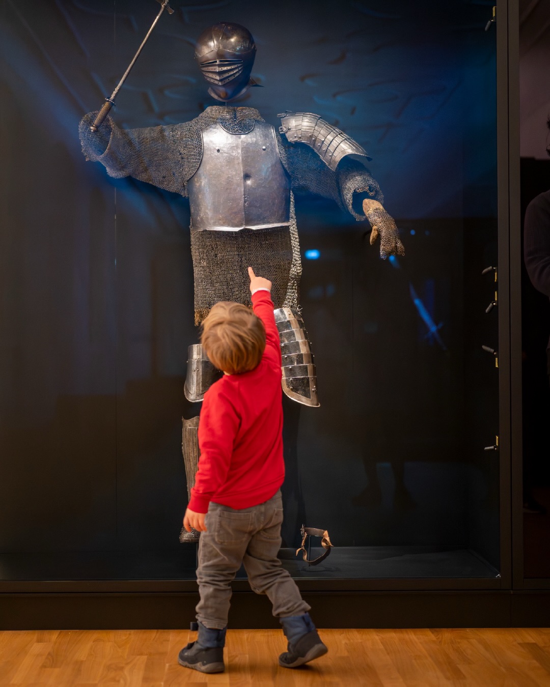 Dieser Alternativtext wurde KI-generiert: Eine Person zeigt auf eine Ritterrüstung in einer Vitrine. Das Bild zeigt Museumsexponate und einen Besucherausblick.