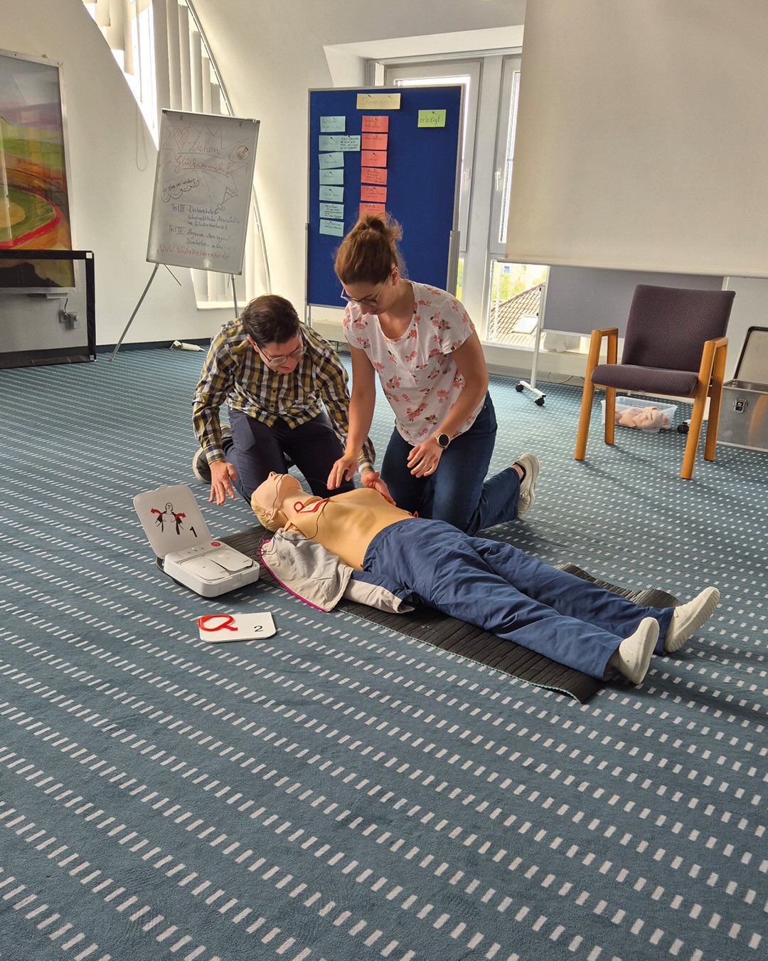 Zwei Personen knien auf dem Teppichboden neben einer liegenden Reanimationspuppe. Eine Person führt Herzdruckmassage durch, während die andere Person daneben kniet und beobachtet. Neben dem Kopf der Puppe liegt ein offener Defibrillator mit Elektroden auf dem Boden. Im Hintergrund sind ein Whiteboard und eine Pinnwand zu sehen. (KI-Alternativtext)