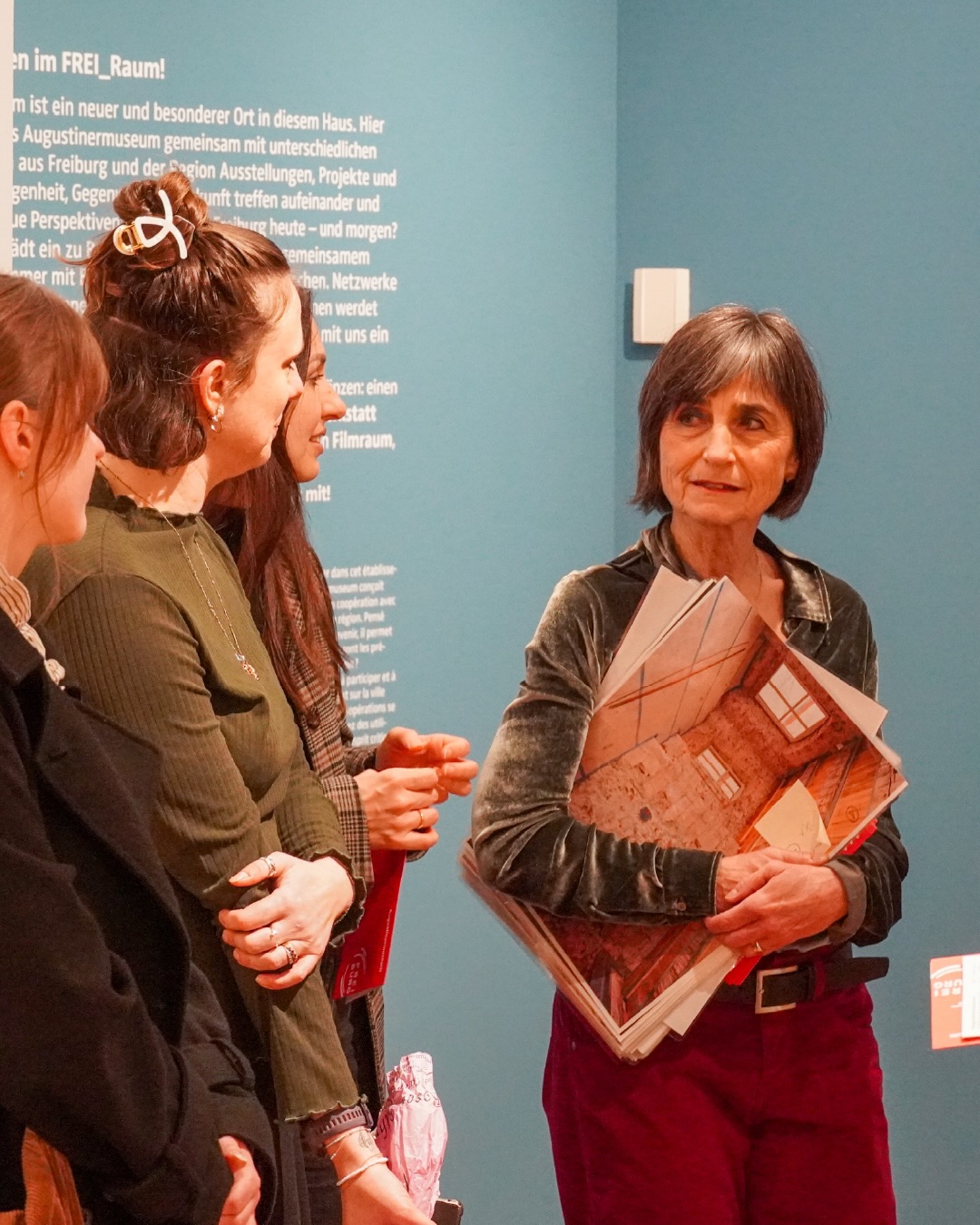 Dieser Alternativtext wurde KI-generiert: Eine farbige Innenaufnahme zeigt drei Personen, die in einem Raum miteinander interagieren. Zwei Personen schauen auf etwas in der Hand einer dritten Person, die Dokumente hält.