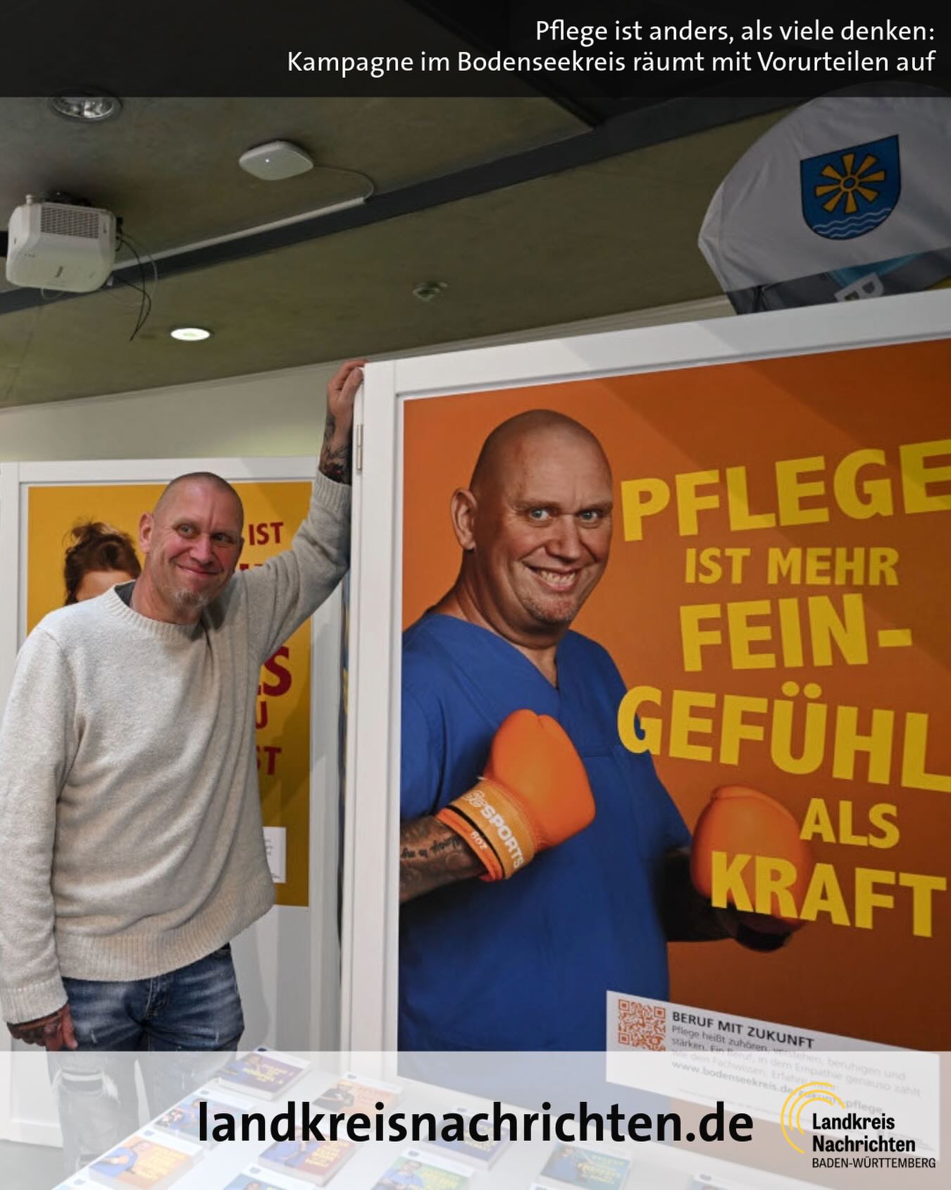 Dieser Alternativtext wurde KI-generiert: Zwei Personen stehen vor einem farbenfrohen Plakat mit Aufschrift. Im Hintergrund sind weitere Personen und ein Raum zu sehen.