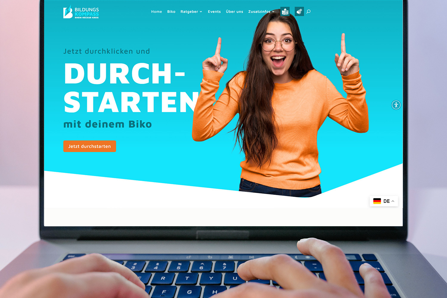 Laptopbildschirm mit der Website des Bildungskompass Rhein-Neckar-Kreis. Eine Person in einem orangefarbenen Pullover zeigt mit beiden Zeigefingern nach oben und wirkt begeistert. Der Text lautet 'Jetzt durchklicken und DURCH-STARTEN mit deinem Biko' mit einem Button 'Jetzt durchstarten'. Im Vordergrund tippen Hände auf der Tastatur. (KI-Alternativtext)