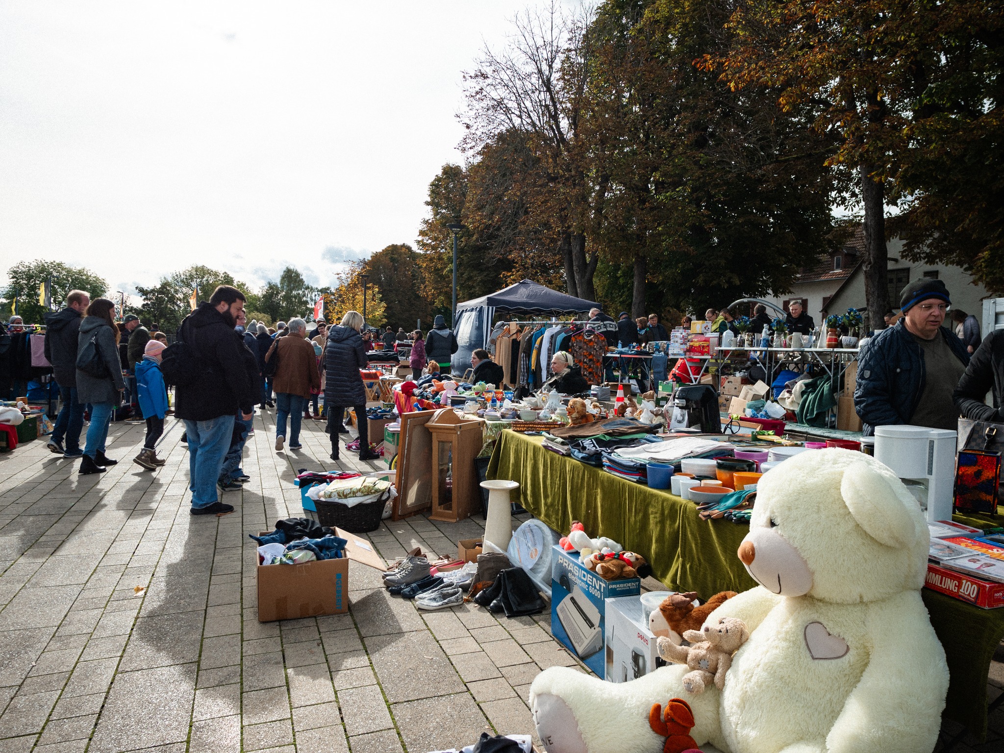 Dieser Alternativtext wurde KI-generiert: Ein belebter Flohmarkt im Freien mit vielen Menschen und Ständen voller verschiedener Waren. Im Vordergrund befindet sich ein großer Teddybär.