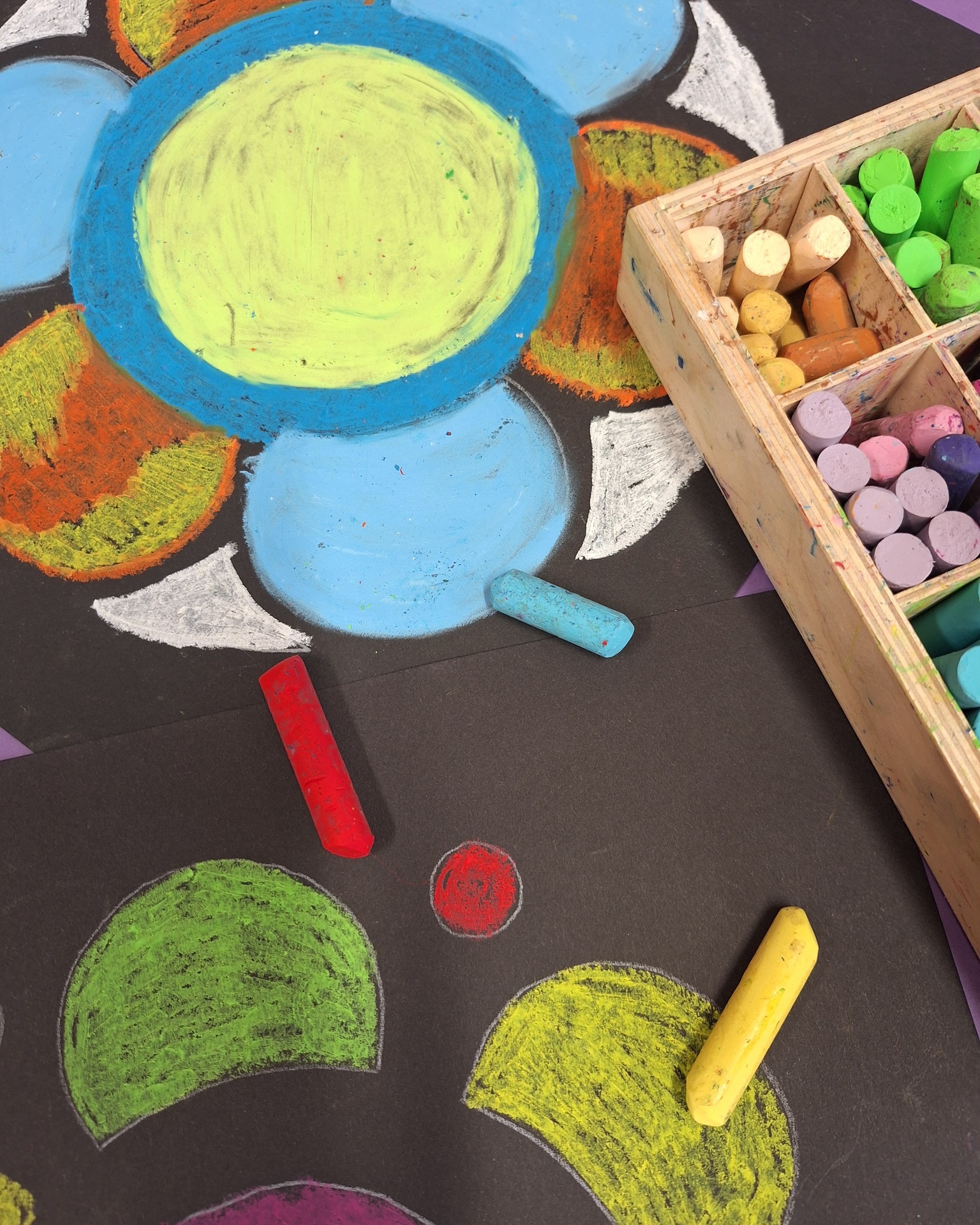 Dieser Alternativtext wurde KI-generiert: Bunte Kreidezeichnungen in Form von Blüten und Kreisen sind auf dunklem Untergrund zu sehen. Neben den Zeichnungen liegt eine Schachtel mit weiteren Kreidestücken.
