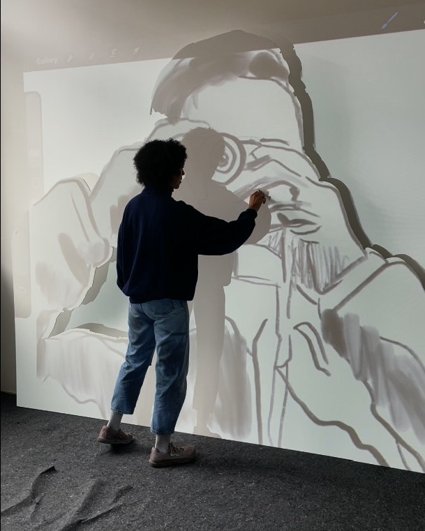 Dieser Alternativtext wurde KI-generiert: Eine Person steht mit dem Rücken zum Betrachter und zeichnet mit einem Stift auf eine große, helle Fläche. Das Bild zeigt eine Skizze menschlicher Formen und Hände.