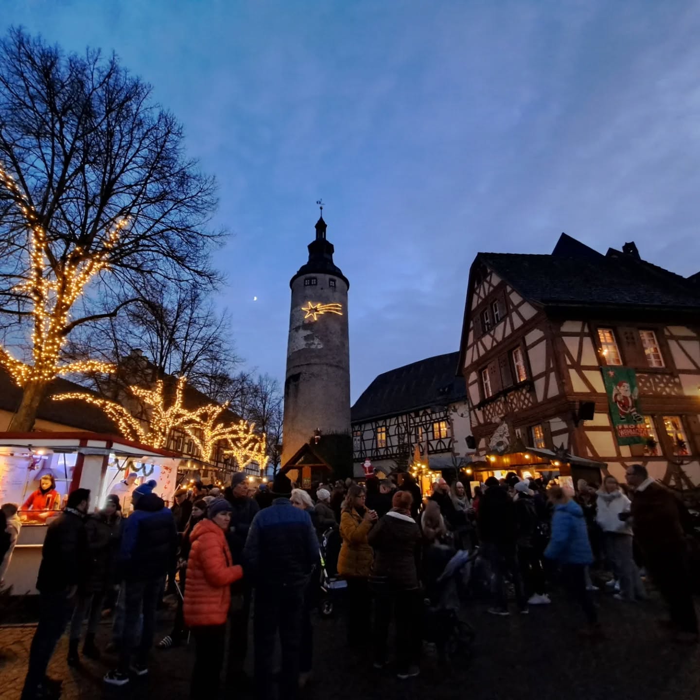 Dieser Alternativtext wurde KI-generiert: Ein Turm und Fachwerkhäuser stehen im Hintergrund einer belebten Szene. Lichterketten und Buden deuten auf einen Weihnachtsmarkt hin.