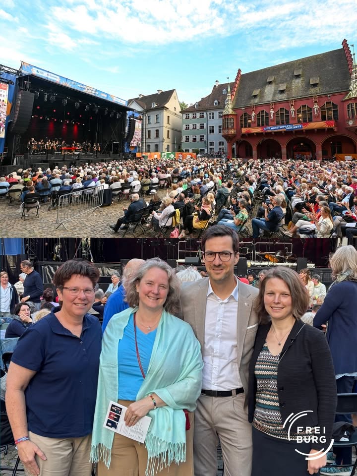 Große Menschenmenge auf einem Freiluftkonzert vor einer Bühne und historischen Gebäuden; im Vordergrund vier Personen, die lächelnd in die Kamera blicken, eine hält ein Programmheft. (KI-Alternativtext)