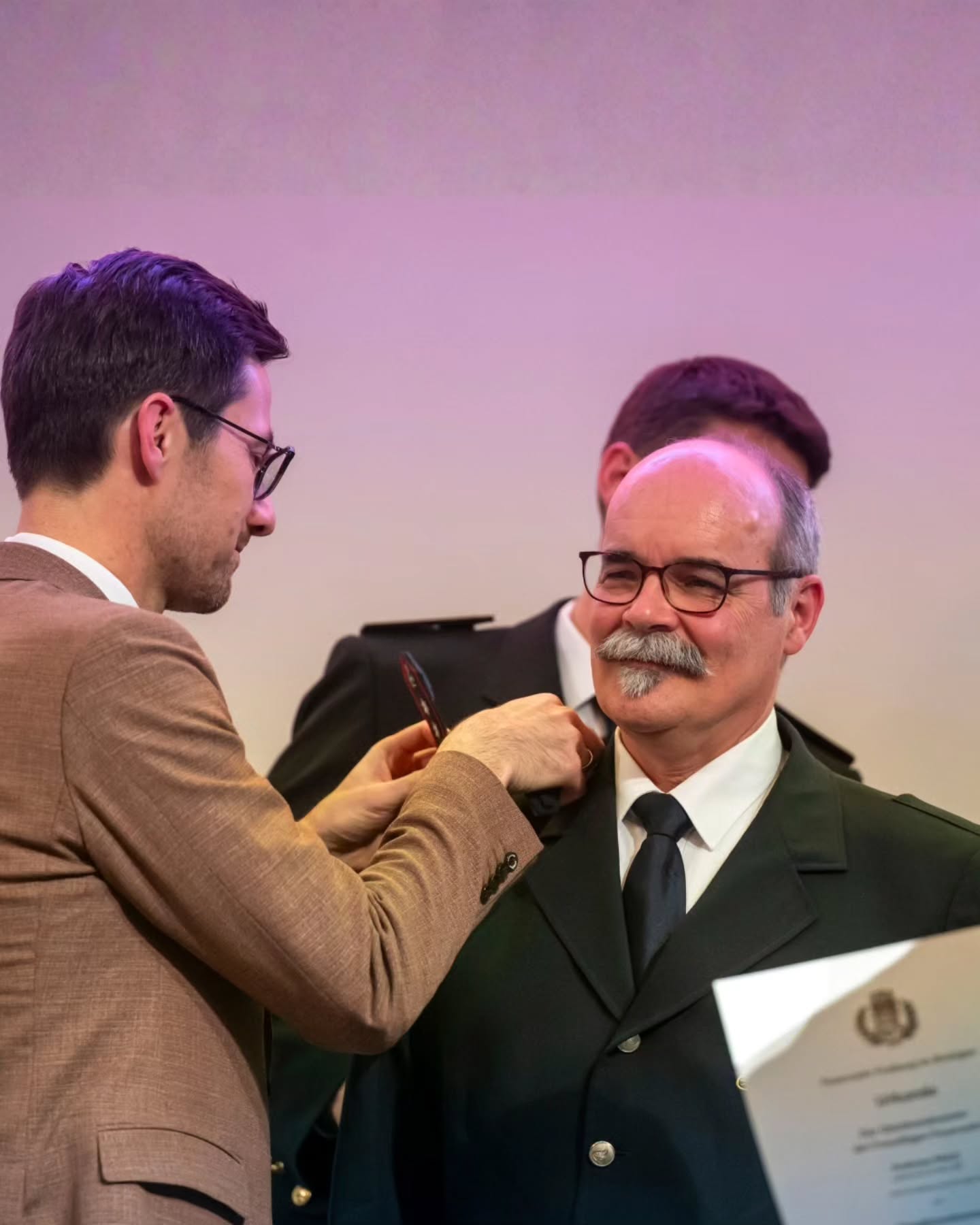 Dieser Alternativtext wurde KI-generiert: Zwei Personen im Gespräch, eine davon überreicht eine Auszeichnung. Im Hintergrund sind weitere Personen und eine helle Wand zu sehen.