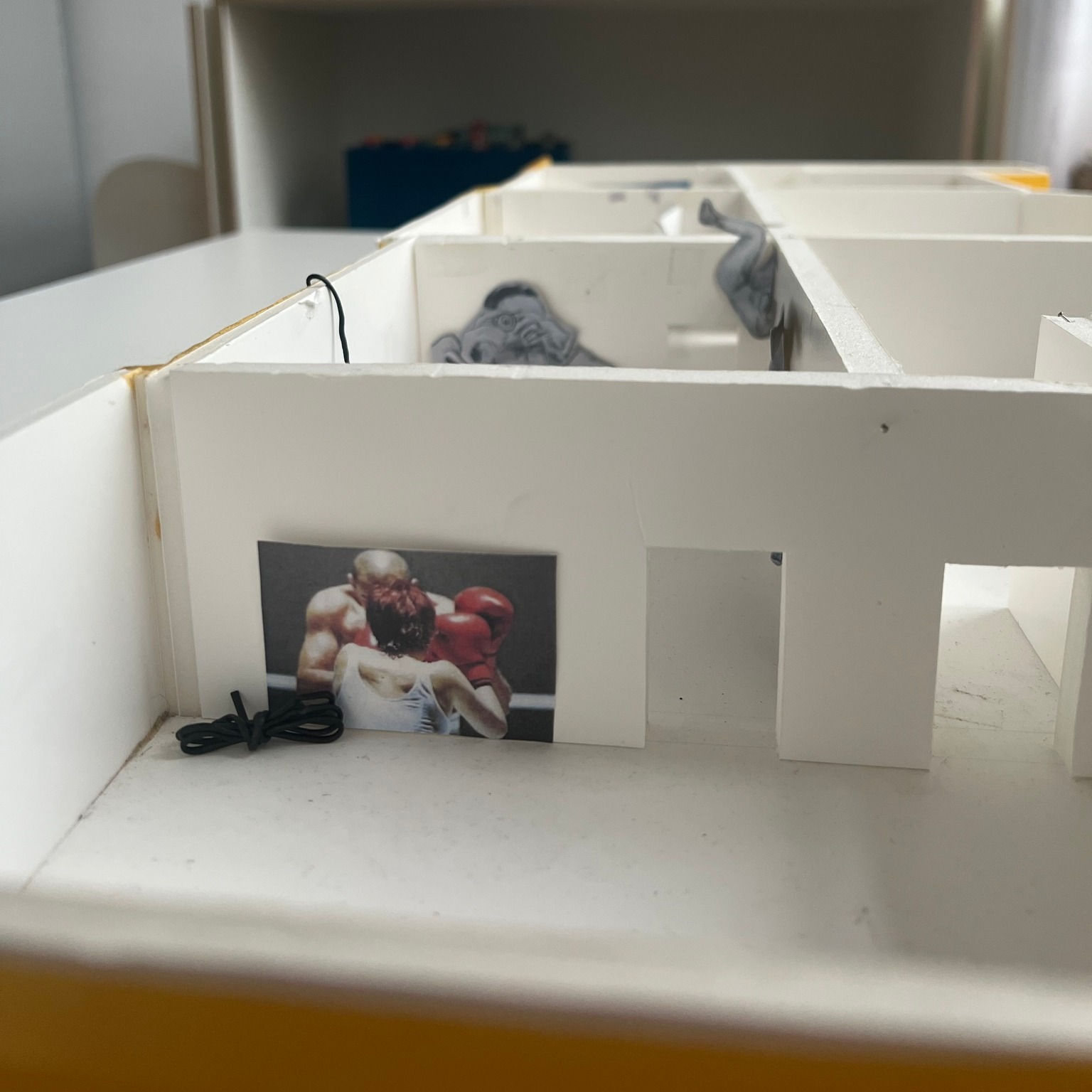 Dieser Alternativtext wurde KI-generiert: Ein Modell aus hellen Holzplatten zeigt einen Innenraum mit Bildern von Boxern. Die Bilder sind in die Struktur des Modells integriert.