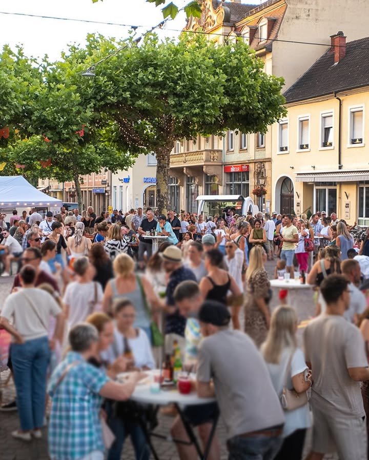 Eine große Menschenmenge auf einer Straße bei einem Straßenfest, wobei Personen im Vordergrund an runden Tischen sitzen und essen. Im Hintergrund stehen weitere Personen, Bäume und Gebäude mit Ladengeschäften. (KI-Alternativtext)