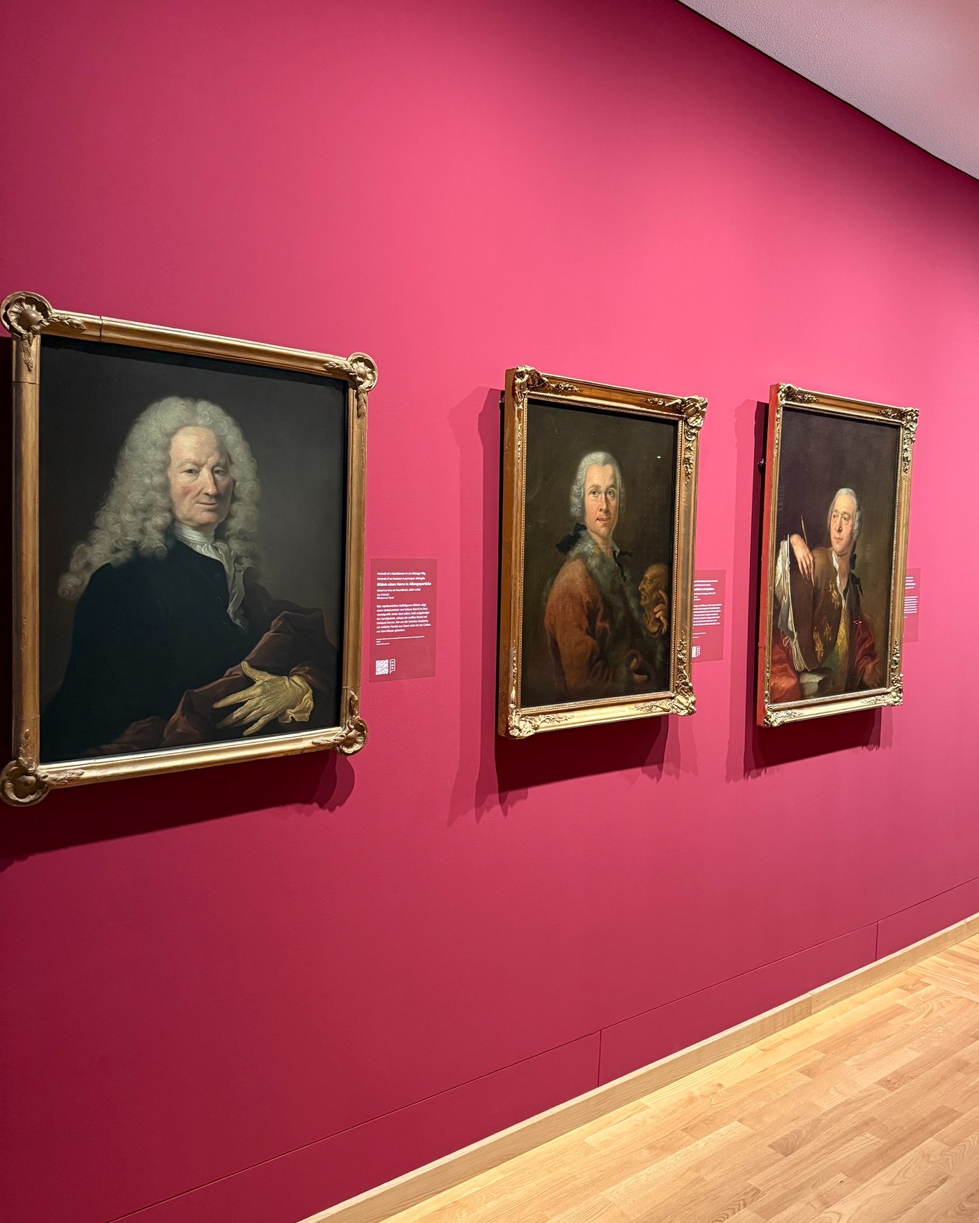 Drei historische Porträts in goldenen Rahmen hängen an einer leuchtend pinken Wand. Jedes Bild zeigt eine Person mit Perücke in historischer Kleidung. Unter den Bildern befinden sich kleine Informationskärtchen. (KI-Alternativtext)