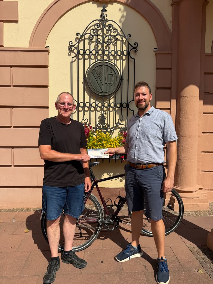 Zwei Personen stehen vor einem Gebäude mit einem verzierten Fenstergitter und halten gemeinsam ein Dokument. Zwischen ihnen steht ein Fahrrad. (KI-Alternativtext)