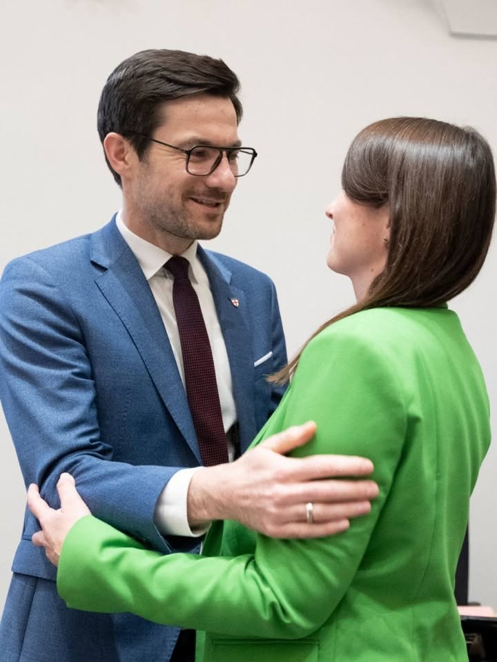 Dieser Alternativtext wurde KI-generiert: Zwei Personen in formeller Kleidung schauen einander an und lächeln. Eine Person umarmt die andere seitlich.