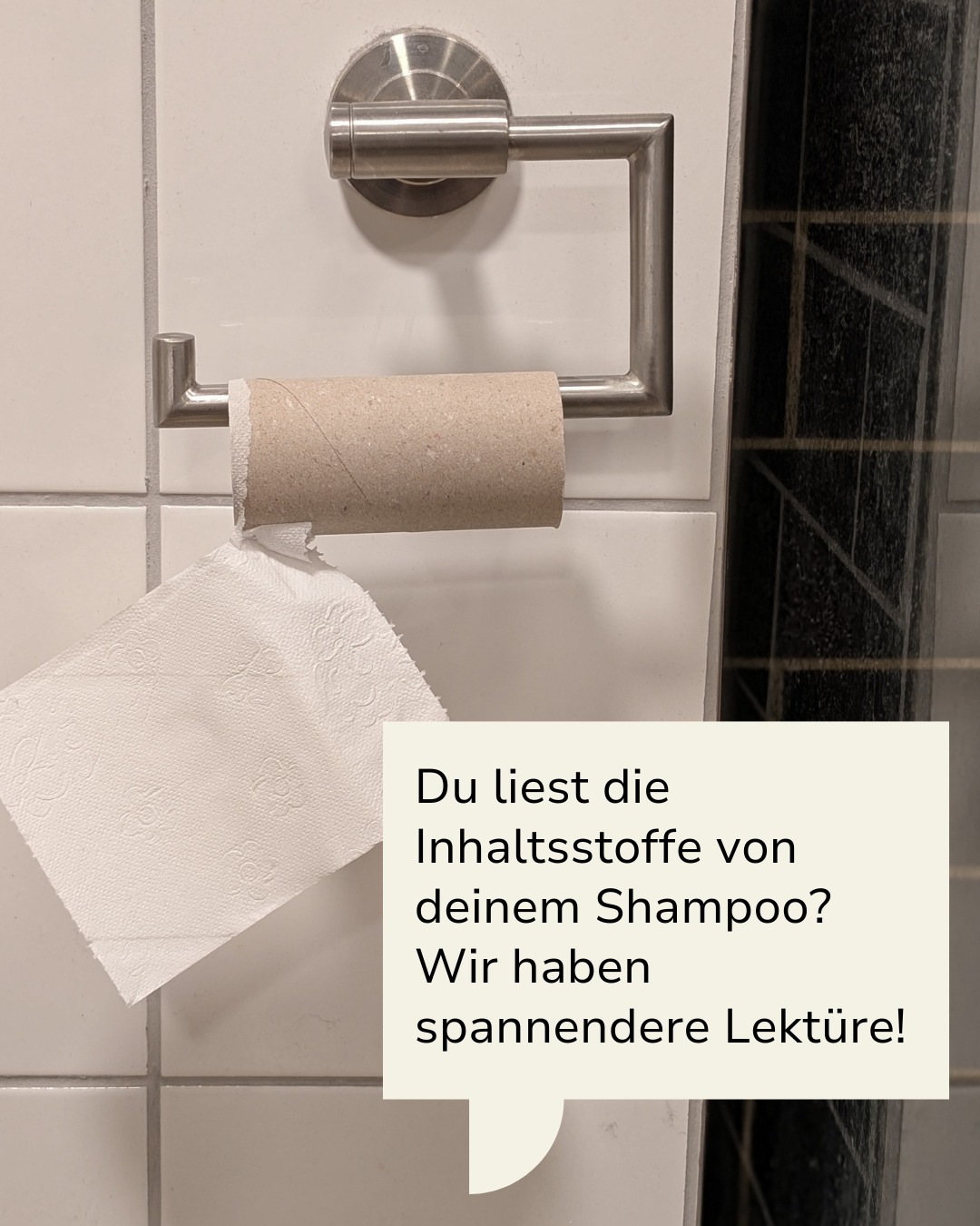 Dieser Alternativtext wurde KI-generiert: Ein Toilettenpapierhalter mit fast leerer Rolle an einer gefliesten Wand. Ein Schild mit Text ist unter dem Papier angebracht.