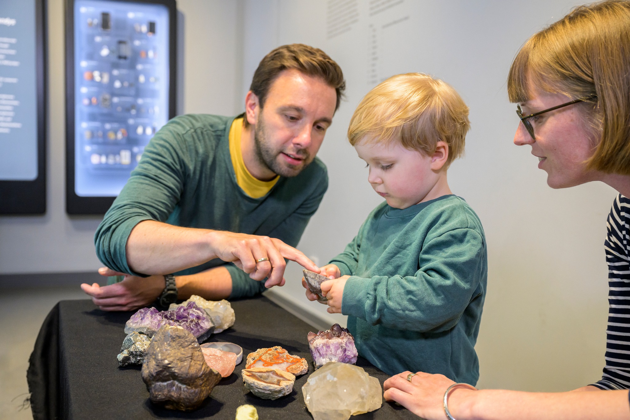 Dieser Alternativtext wurde KI-generiert: Drei Personen betrachten gemeinsam verschiedene Kristalle, die auf einem Tisch ausgestellt sind. Im Hintergrund ist eine Ausstellungswand zu sehen.