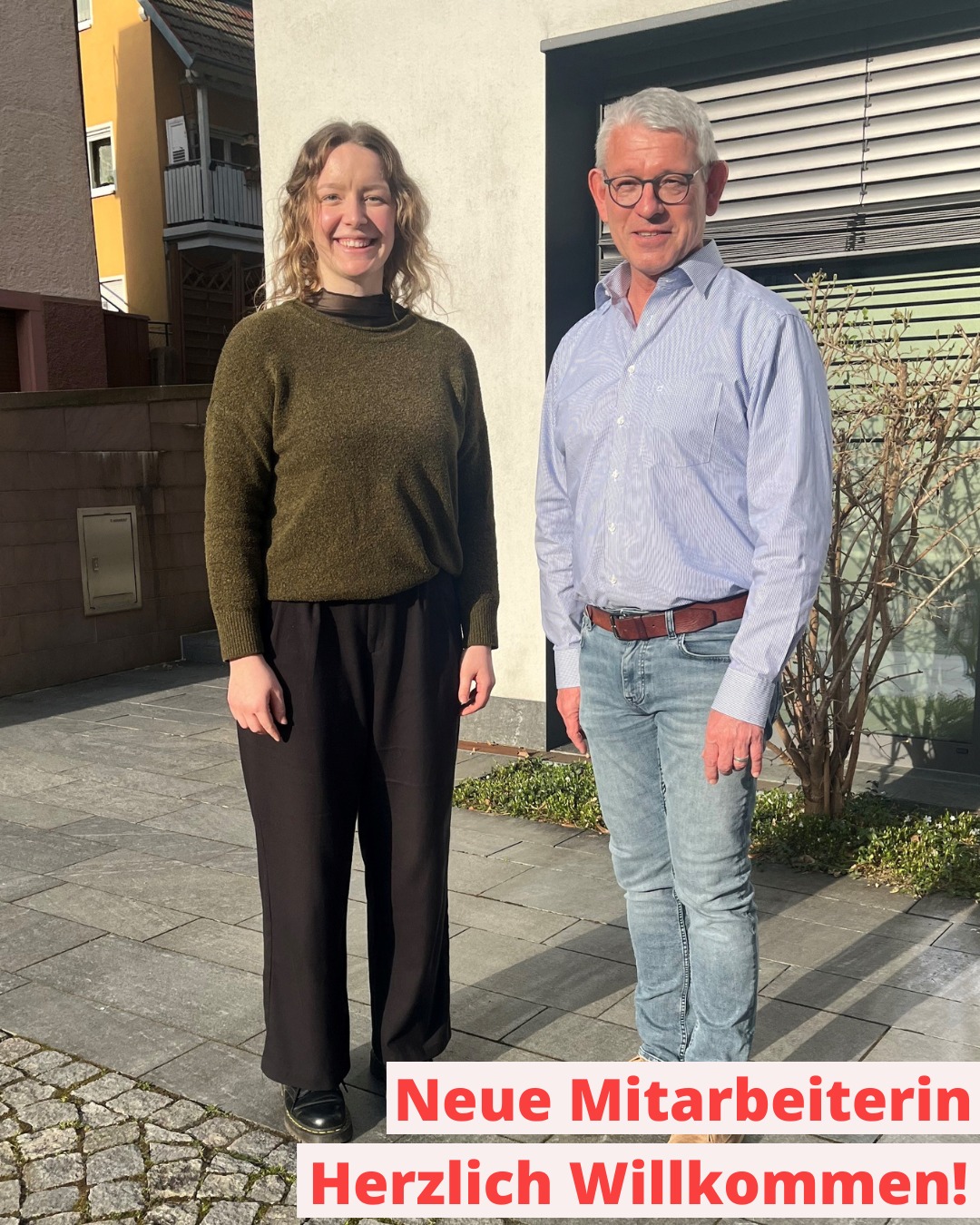 Dieser Alternativtext wurde KI-generiert: Zwei Personen stehen nebeneinander vor einem Gebäude und einem Busch. Ein roter Textblock mit der Aufschrift "Neue Mitarbeiterin Herzlich Willkommen!" befindet sich im Vordergrund.