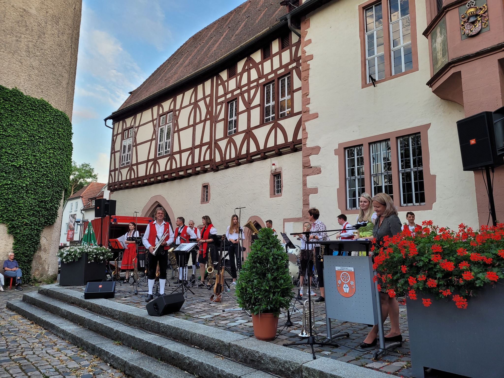 Eine Gruppe von Musikern in traditioneller Kleidung spielt auf einer Bühne vor einem Fachwerkhaus. Eine Person steht am Rednerpult, daneben Blumenkübel und Lautsprecher. Im Hintergrund sitzt ein Zuschauer an einer grün bewachsenen Wand. (KI-Alternativtext)