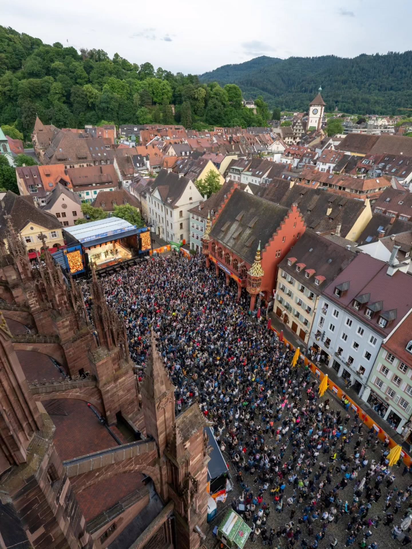 Luftaufnahme eines belebten Marktplatzes in einer historischen Stadt mit dichter Menschenmenge vor einer Bühne, umgeben von alten Gebäuden und einem markanten Kirchturm, im Hintergrund bewaldete Hügel. (KI-Alternativtext)