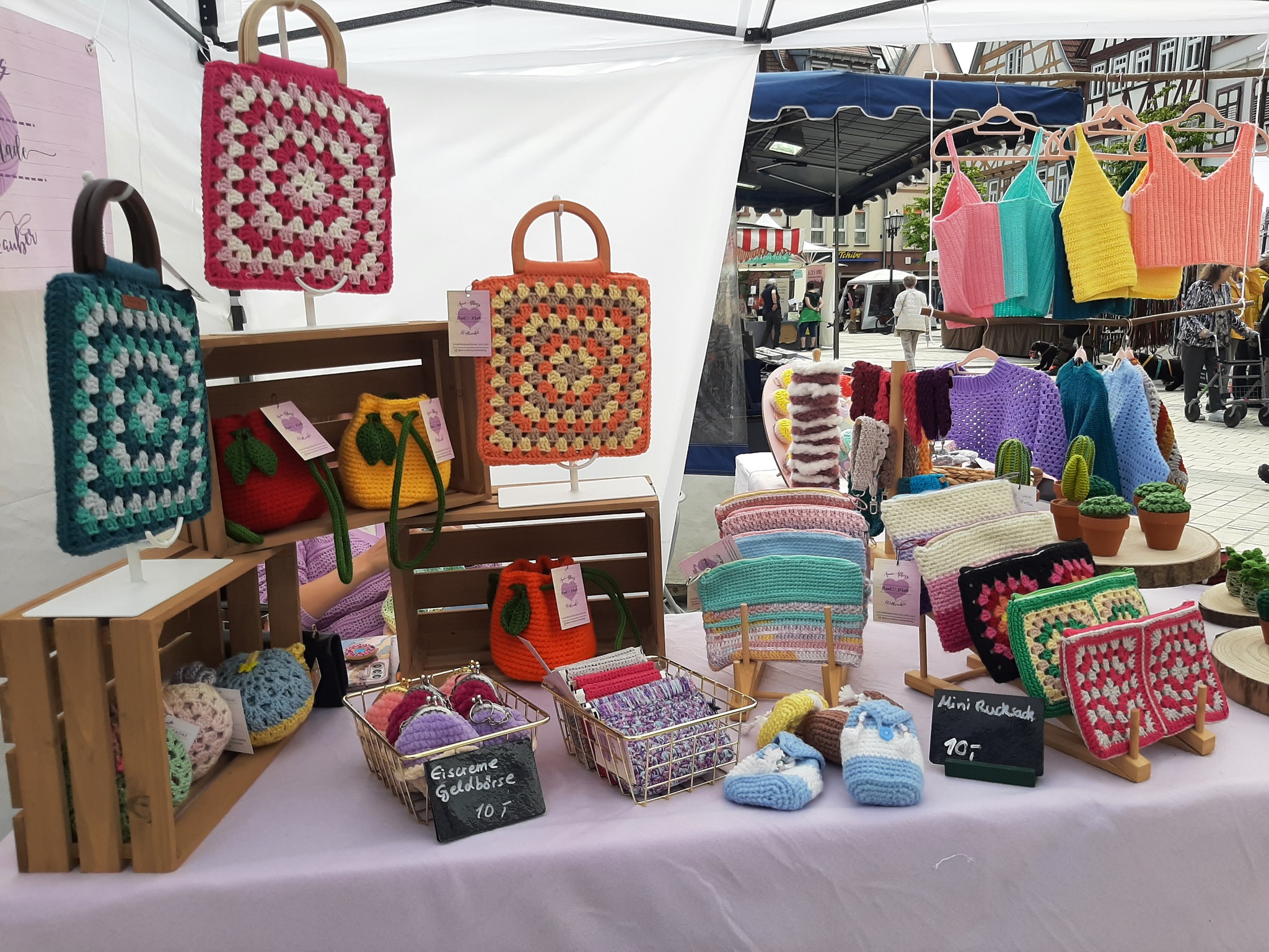 Handgestrickte Taschen, Beutel und Kleidung werden auf einem Marktstand ausgestellt. Diverse bunte Farben und Muster sind zu sehen.