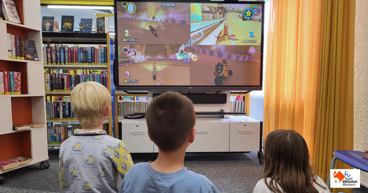 Drei Kinder schauen von hinten auf einen großen Bildschirm, auf dem ein Rennspiel mit vier Spielern läuft. Im Hintergrund sind Bücherregale und ein Fenster mit gelben Vorhängen zu sehen. Unten rechts ist das Logo der Stadtbibliothek Wiesloch. (KI-Alternativtext)