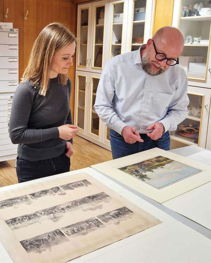 Zwei Personen stehen an einem Tisch und betrachten darauf ausgelegte Kunstwerke. Im Vordergrund liegt eine lange, mehrteilige Schwarz-Weiß-Zeichnung, daneben ein kleineres farbiges Bild. Im Hintergrund stehen hölzerne Vitrinen mit Glasfronten. (KI-Alternativtext)