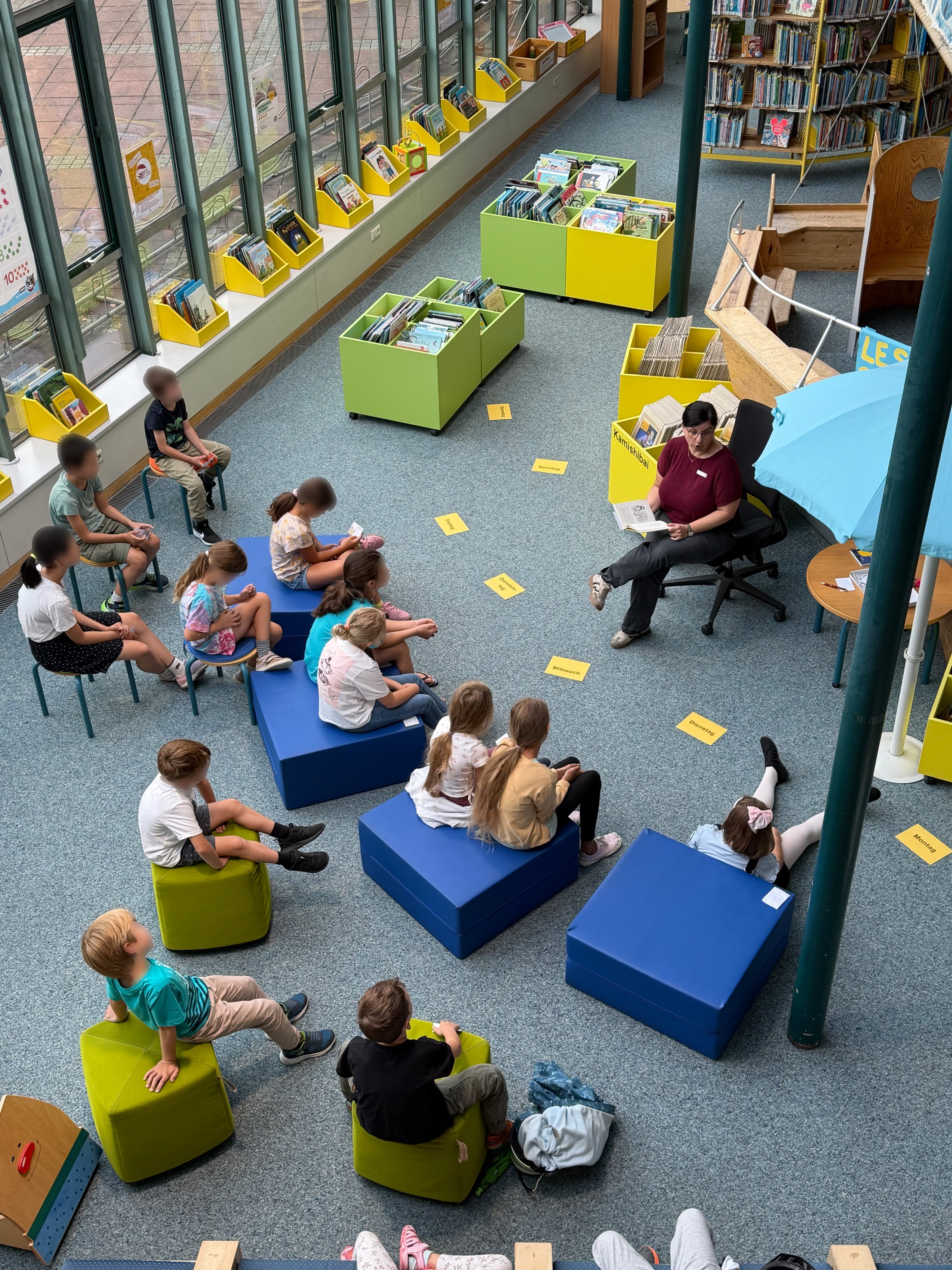 Eine Gruppe von Kindern sitzt im Halbkreis auf blauen und grünen Sitzkissen sowie kleinen Stühlen in einem Bibliotheksbereich. Eine Person sitzt ihnen gegenüber auf einem Bürostuhl und liest aus einem Buch vor, während auf dem Teppichboden gelbe Karten mit den Wochentagen Montag bis Sonntag ausgelegt sind. Im Hintergrund stehen bunte Kisten und Regale voller Bücher. (KI-Alternativtext)