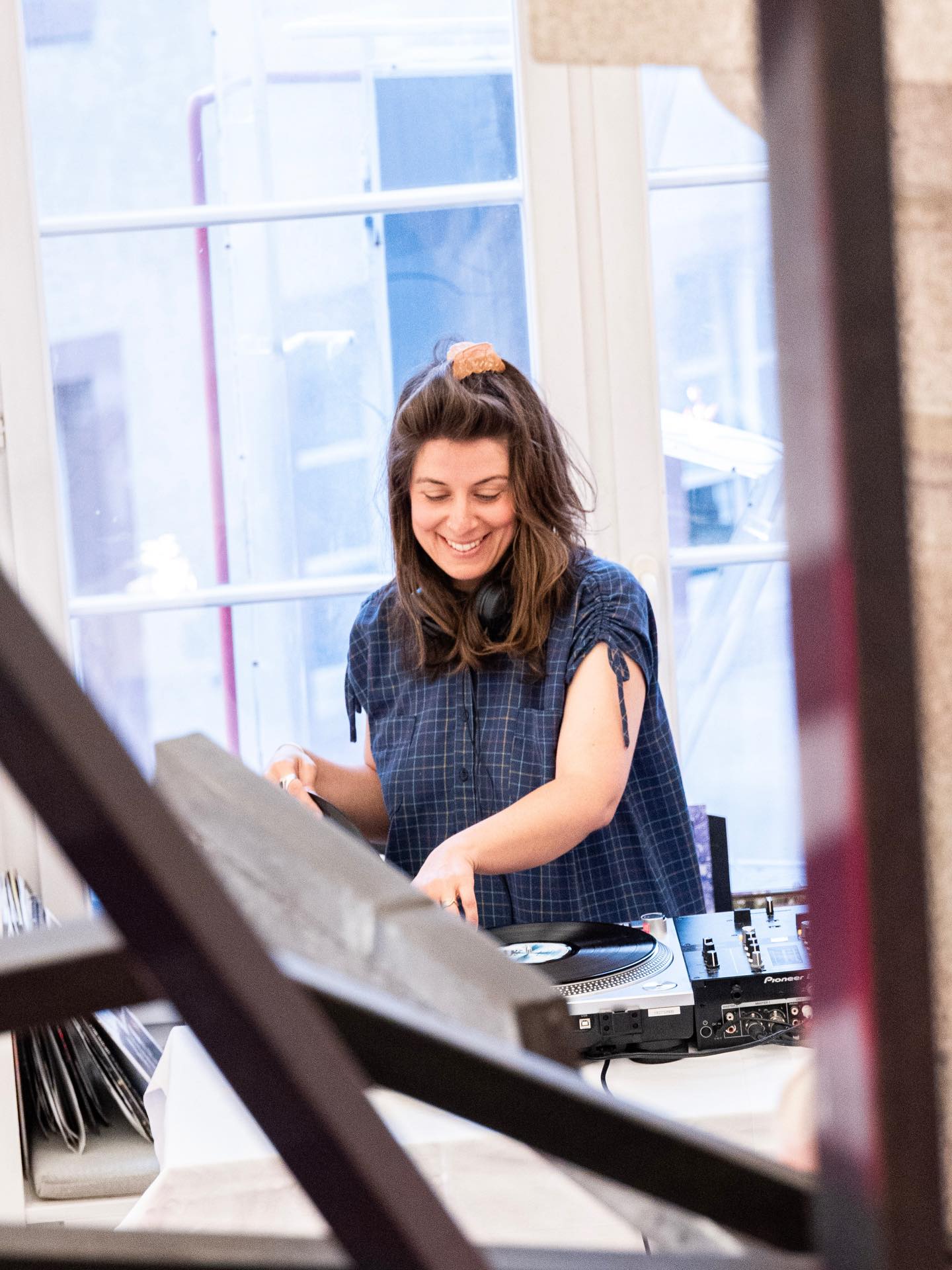 Dieser Alternativtext wurde KI-generiert: Eine Person steht hinter einem DJ-Pult mit Plattenspielern und mischt Musik. Das Bild ist farbig und zeigt eine Innenaufnahme mit einem Fenster im Hintergrund.